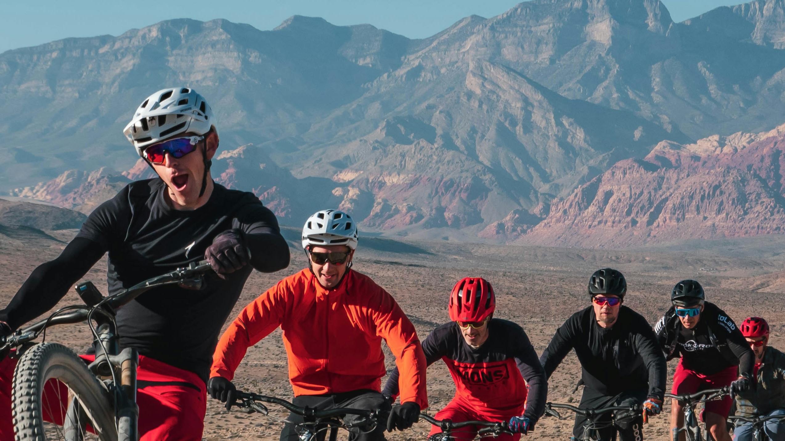 Specialized General Manager Sam Benedict mountain biking and leading a group of five other friends on mountain bikes down a desert path with beautiful, red-tinged mountains in the distance.
