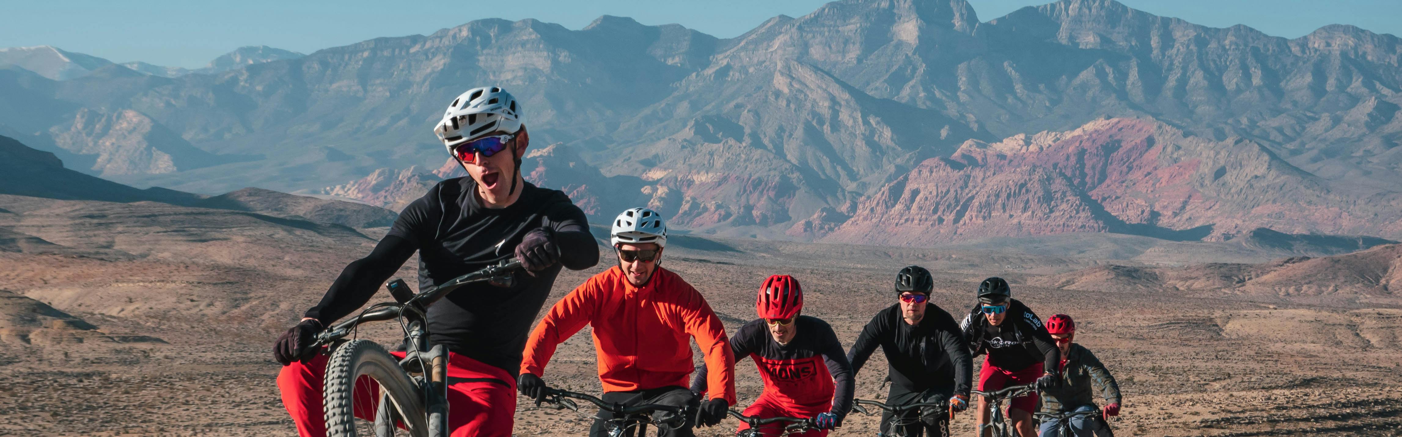 Specialized General Manager Sam Benedict mountain biking and leading a group of five other friends on mountain bikes down a desert path with beautiful, red-tinged mountains in the distance.