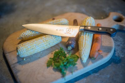 The Mercer Culinary Renaissance Forged Chef's Knife, 8 Inch lying on top of some corn on a cutting board. 