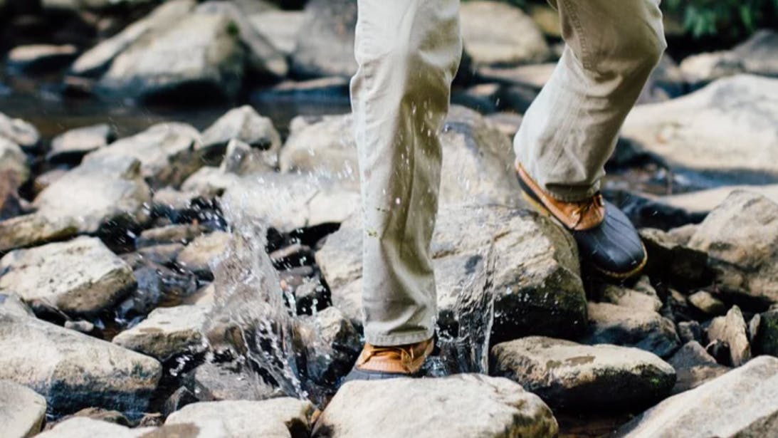 Someone in pants and duck boots crosses a stream. One of their feet splashes in the water.