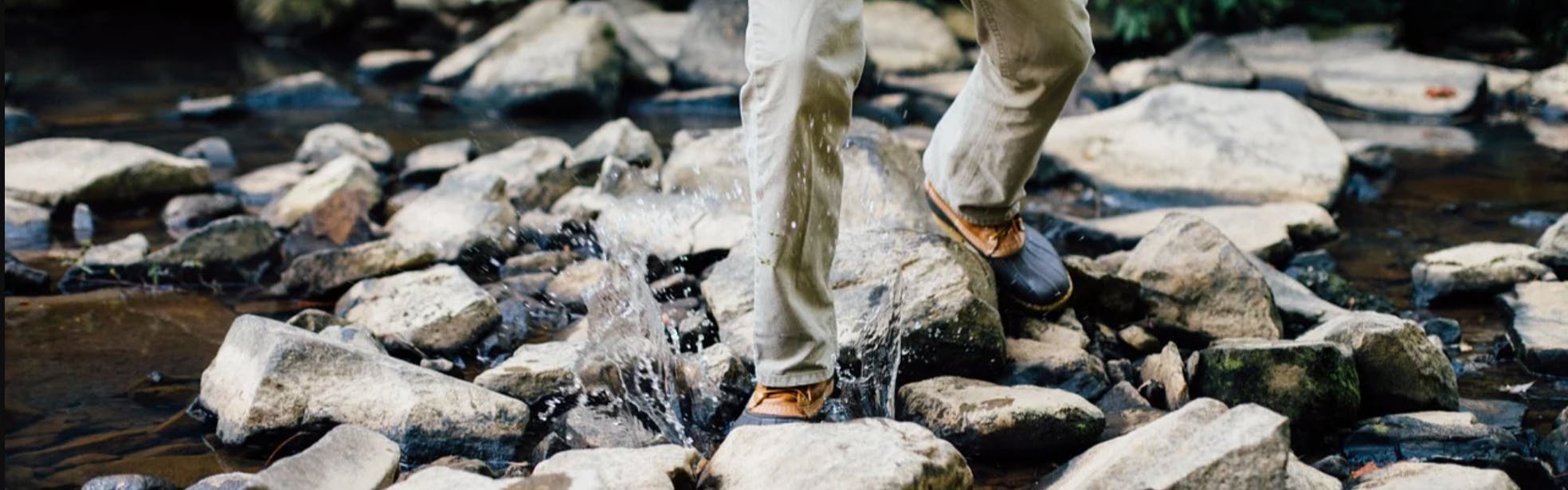 Someone in pants and duck boots crosses a stream. One of their feet splashes in the water.