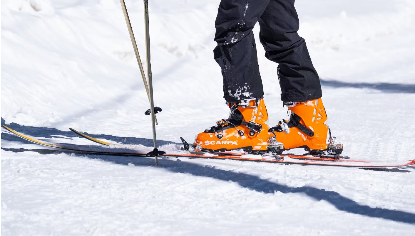 A skier standing on a ski run. He has orange ski boots. 