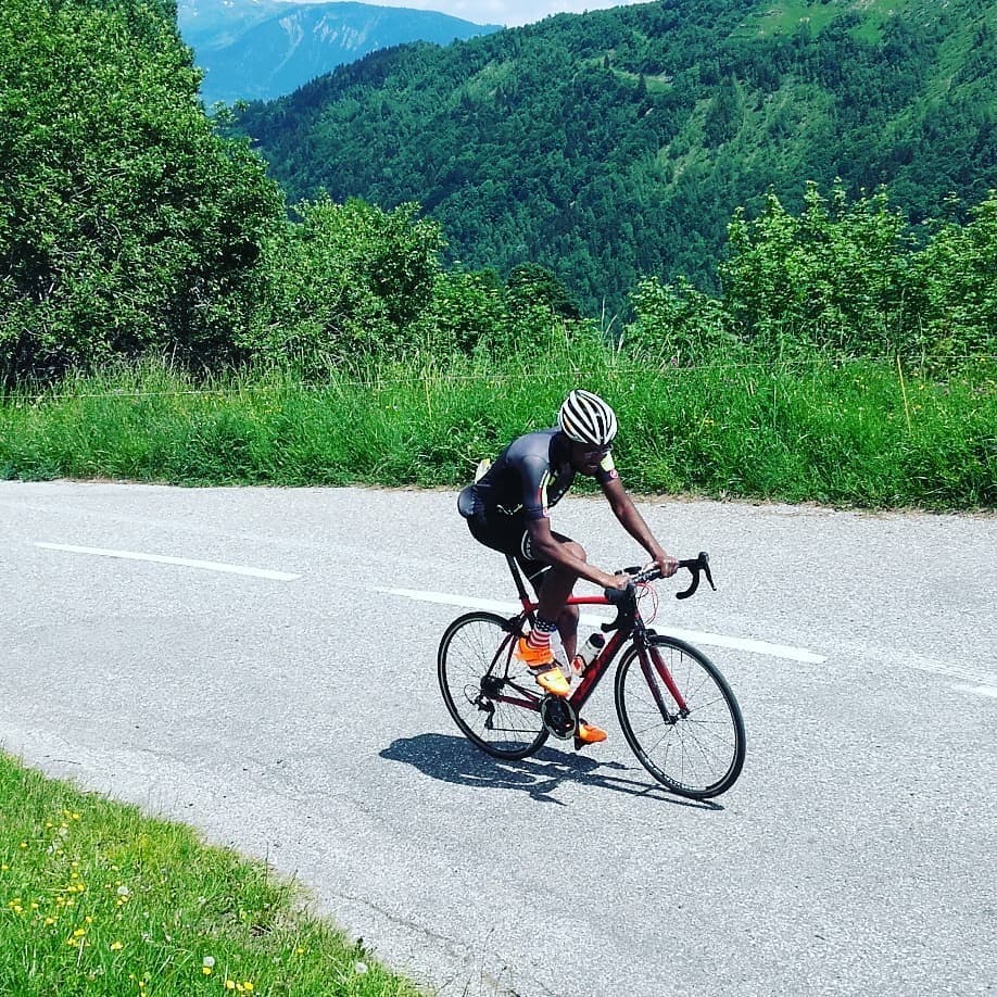 A cyclist climbs a steep looking hill on a road bike. There are a lot of trees and grasses in the background and some mountains in the distance.
