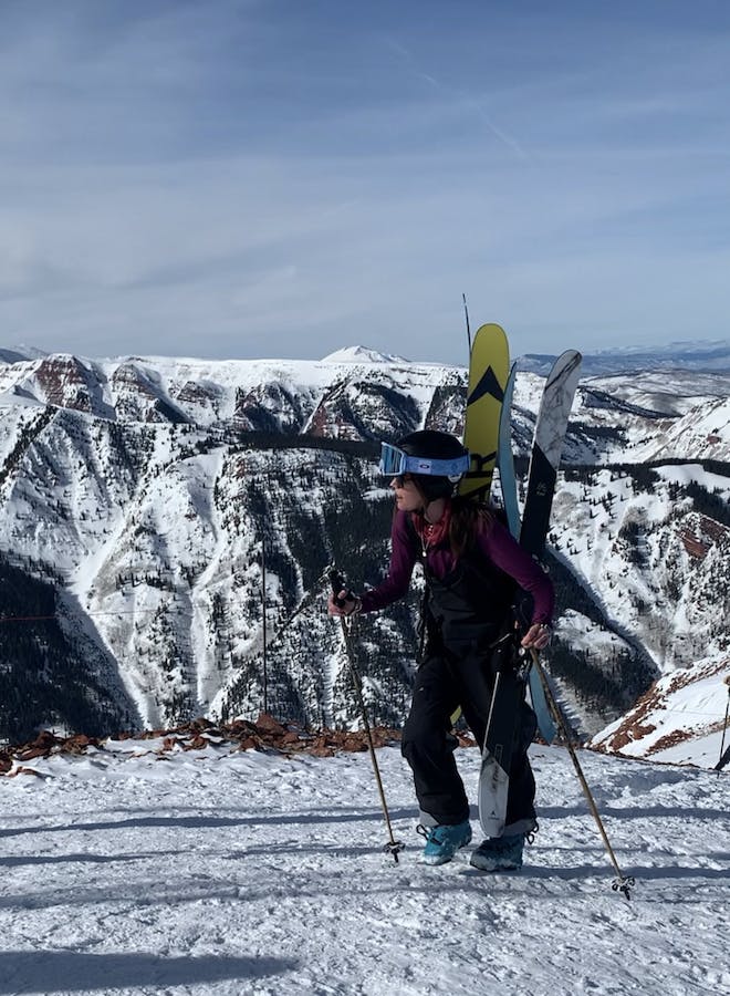 A skier standing at the top of a snowy mountain with her skis on her backpack. 