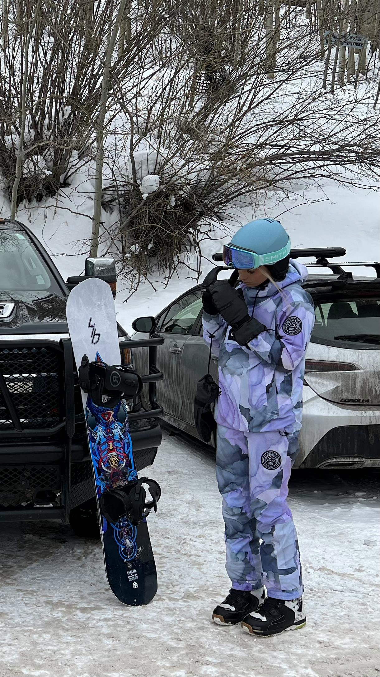 A snowboarder standing in the parking lot looking at the  Lib Tech T.Rice Pro Snowboard · 2023 as it leans against a car. 