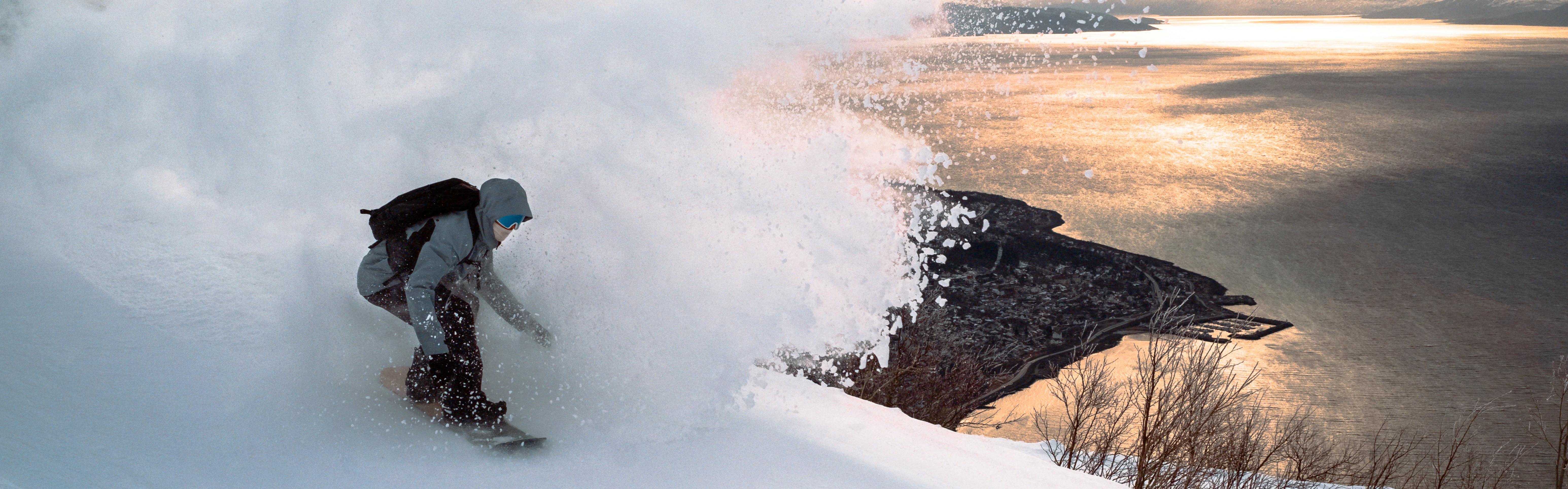 A snowboarder rides down a hill, sending off a big plume of powder behind them. In the distance, a large body of water reflects warm sunlight. 