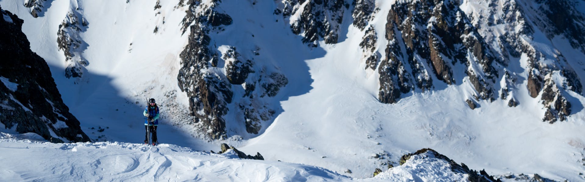 A skier stands on a snowy ridge with a rocky, snowy mountain behind him.