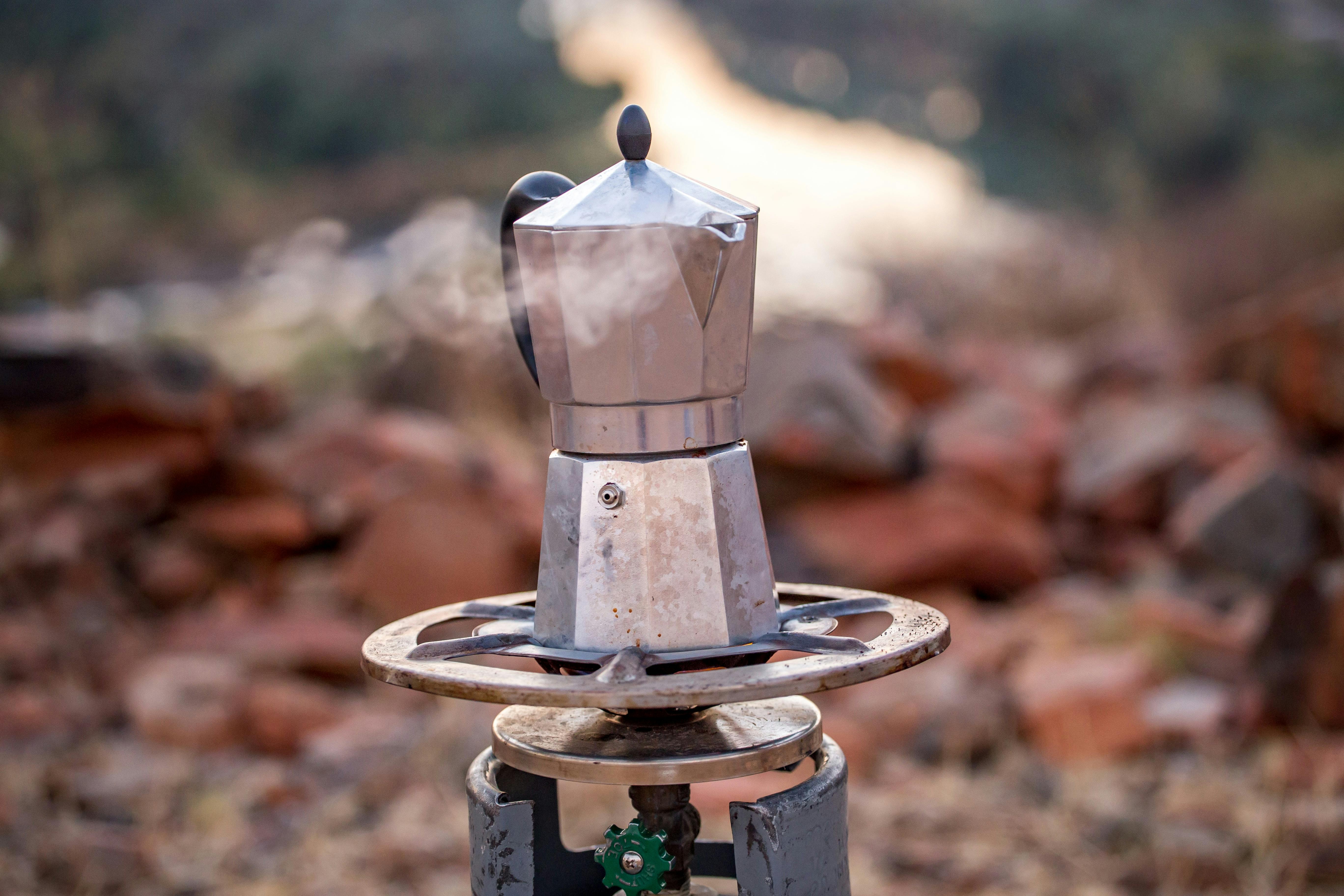 A moka pot coffee maker sits on a camp stove and releases steam