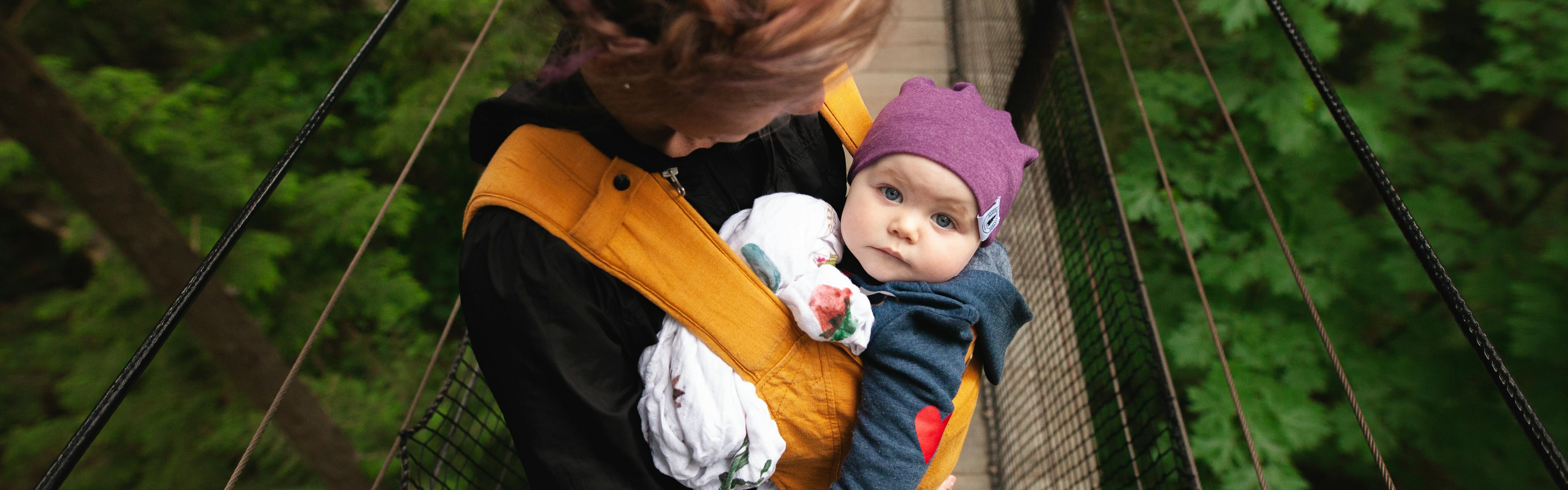 A woman standing on a bridge with a baby in an orange baby carrier. She is looking down at the baby and the baby is looking up at the camera. 
