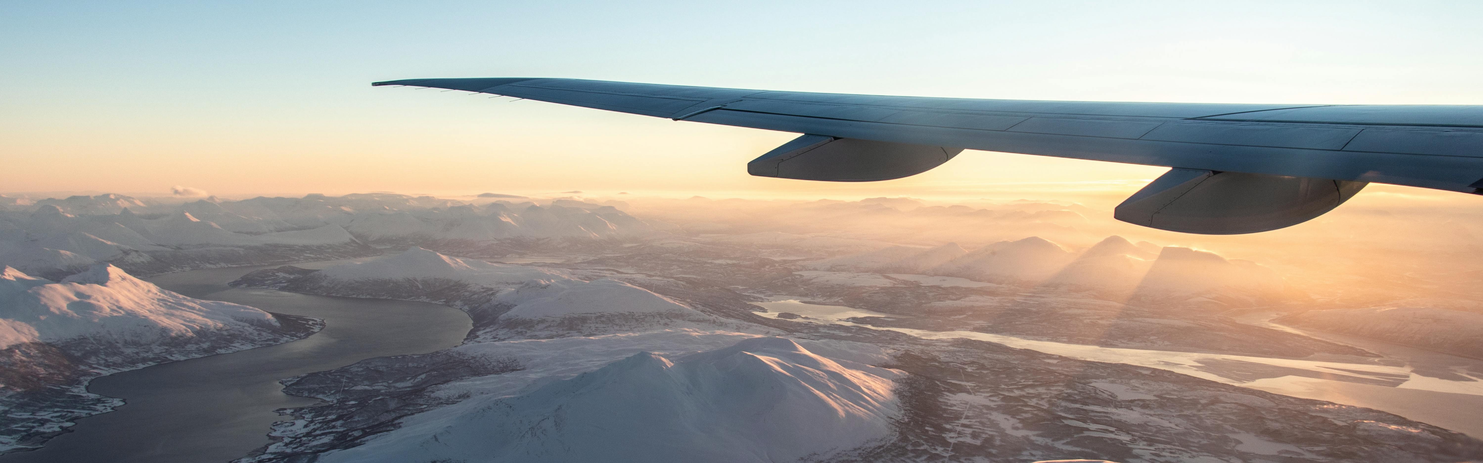 Someone looks at the wing of an airplane through the window. The sun is setting on a snowy landscape on the ground below. 