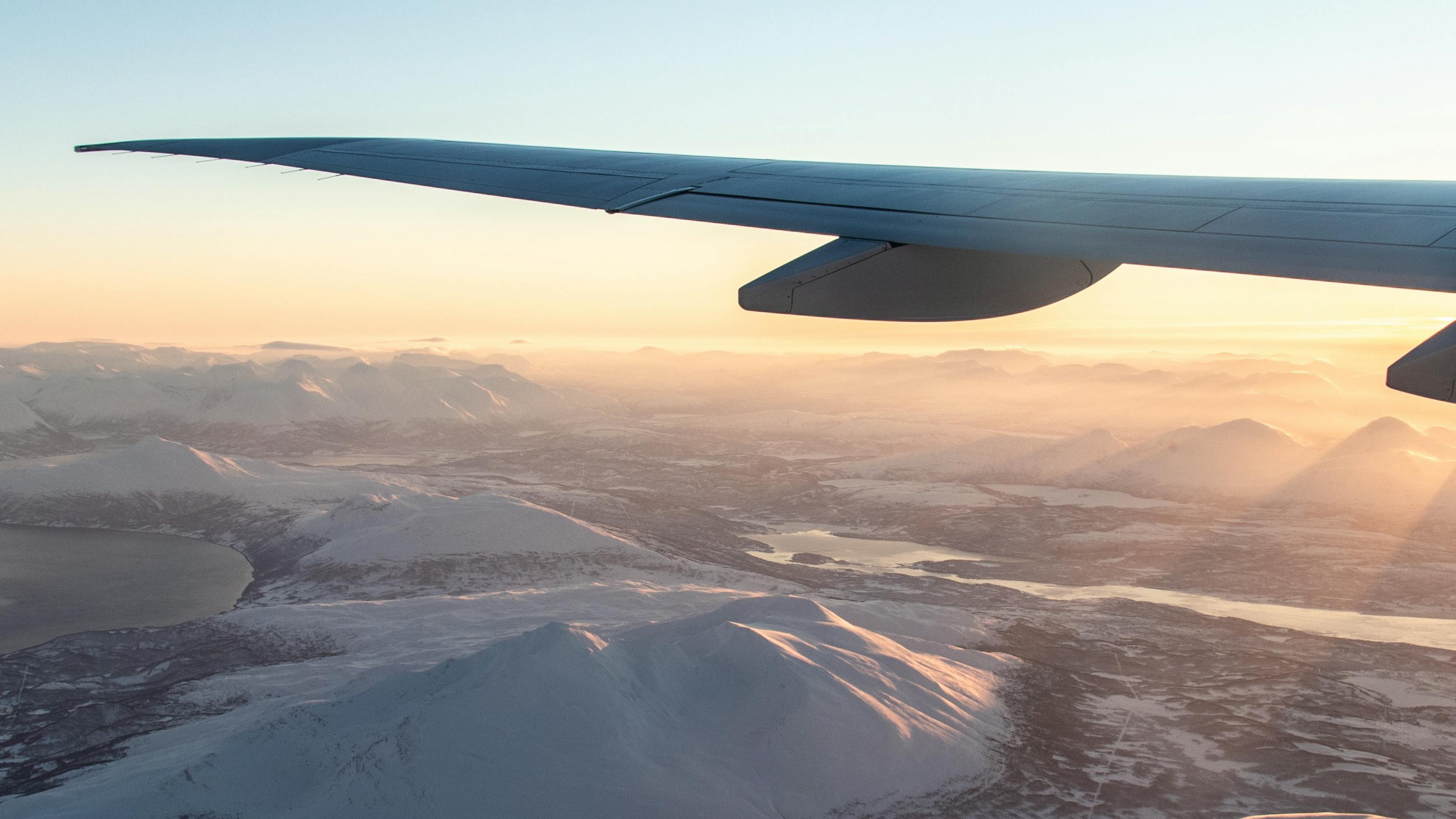 Someone looks at the wing of an airplane through the window. The sun is setting on a snowy landscape on the ground below. 