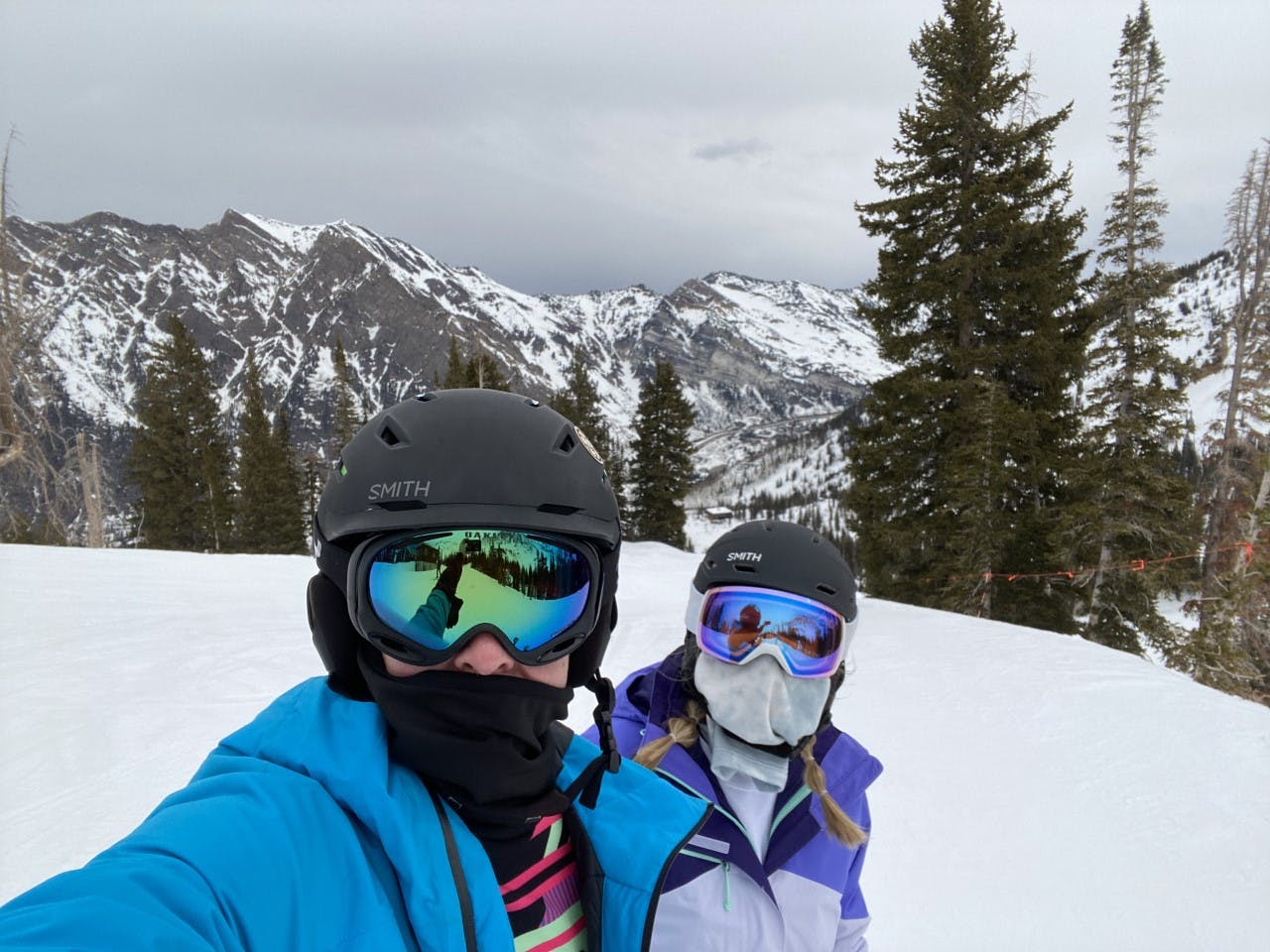 Two skiers both with helmets and goggles on pose for a photo with snowy mountains in the background. 