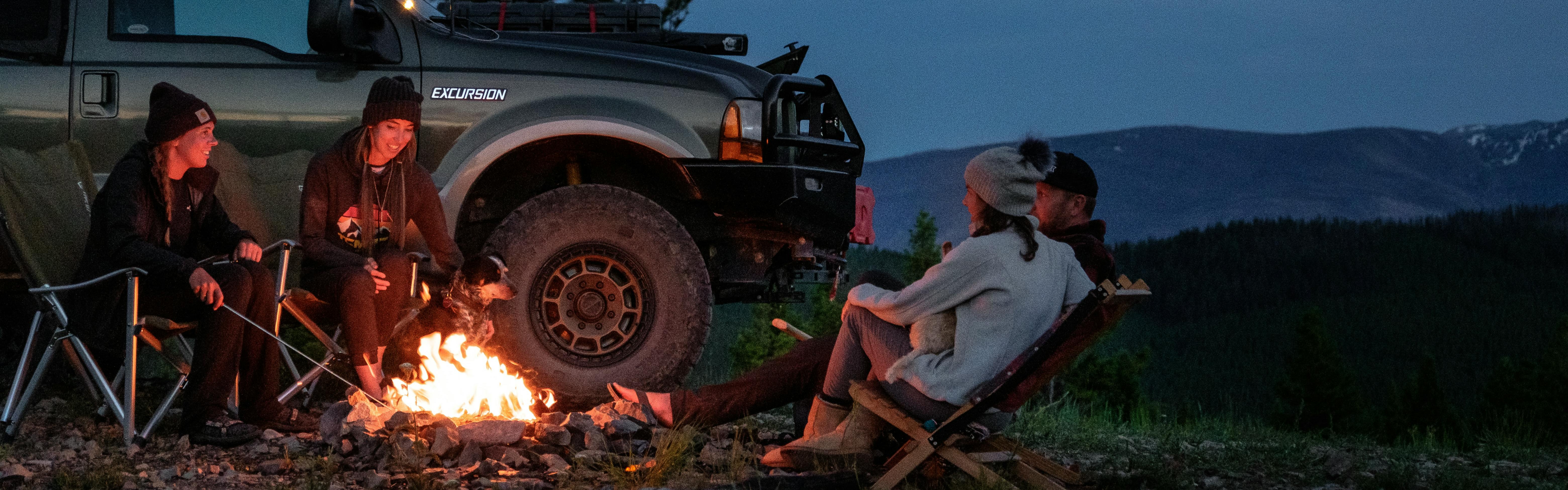 Four people sit around a campfire next to their car at night.