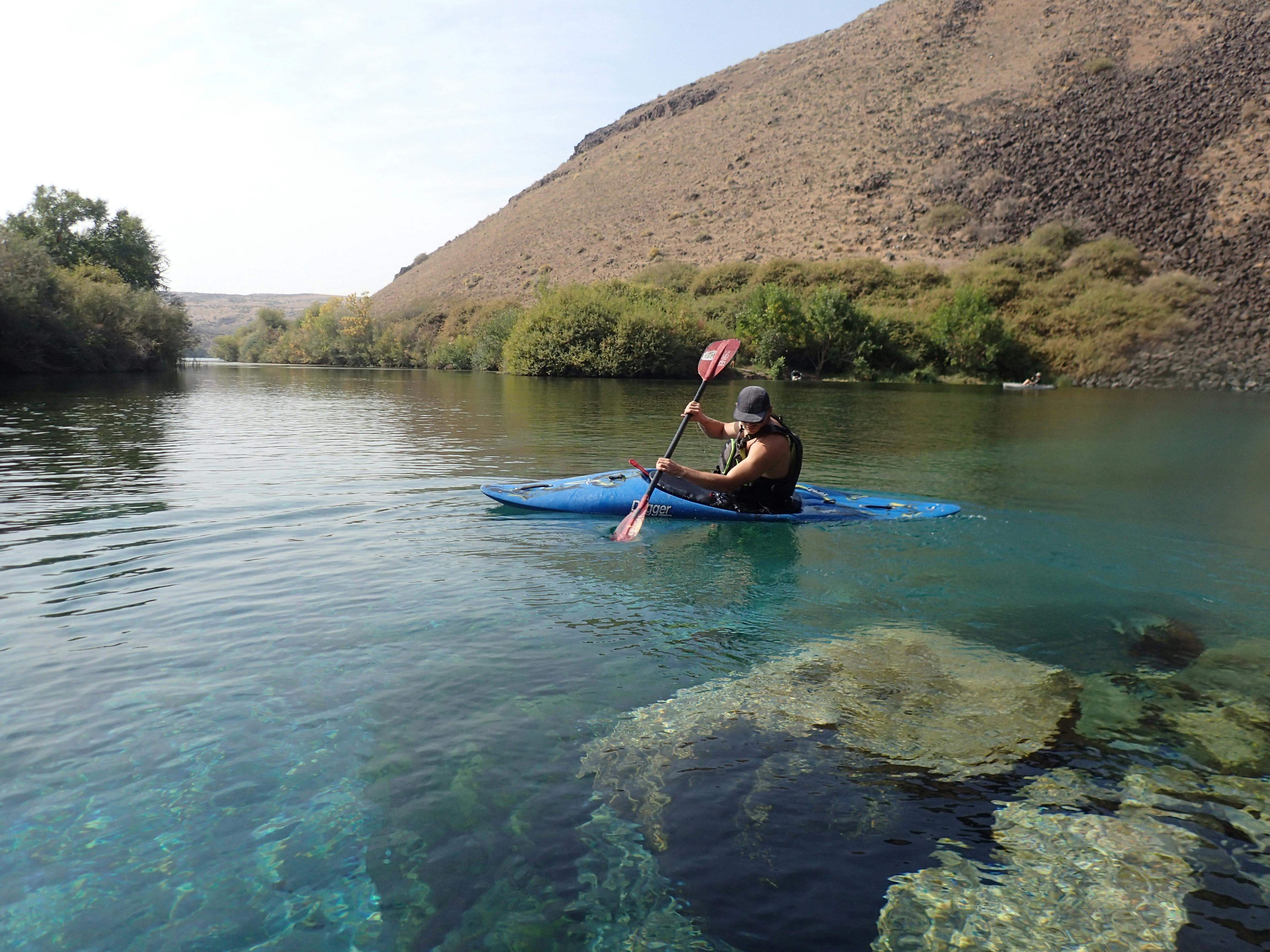 Someone paddles in a blue kayak on clear blue water with brown hills in the background.