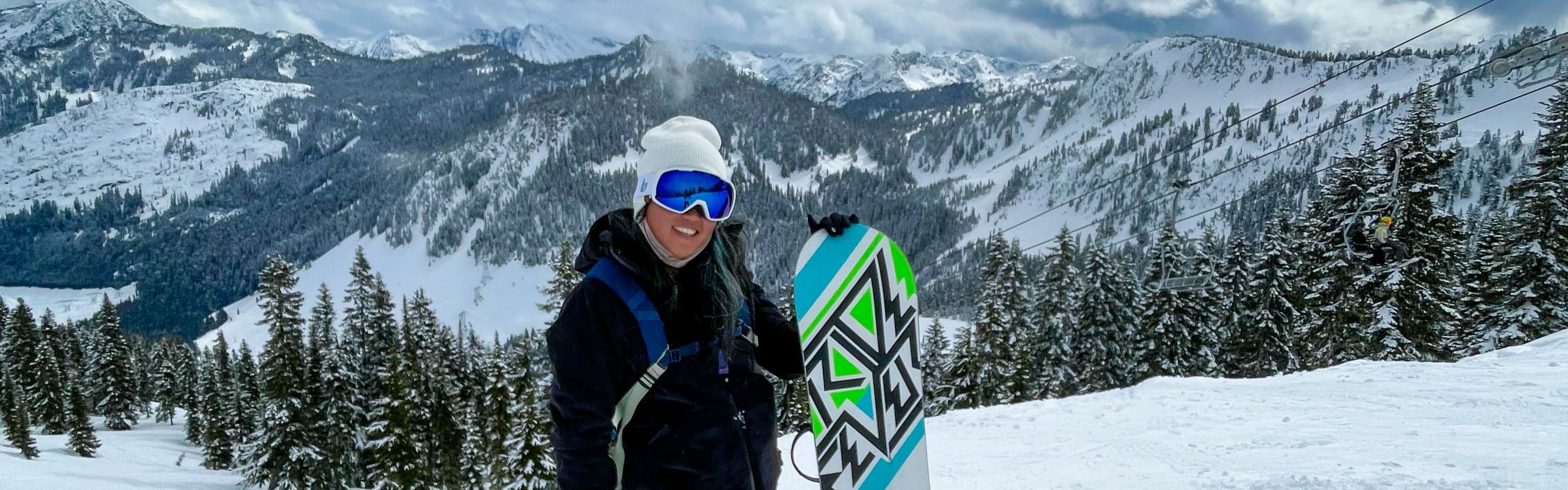 A woman stands holding her snowboard and smiles for the camera. Snowy, tree-covered mountains ruse behind her. 
