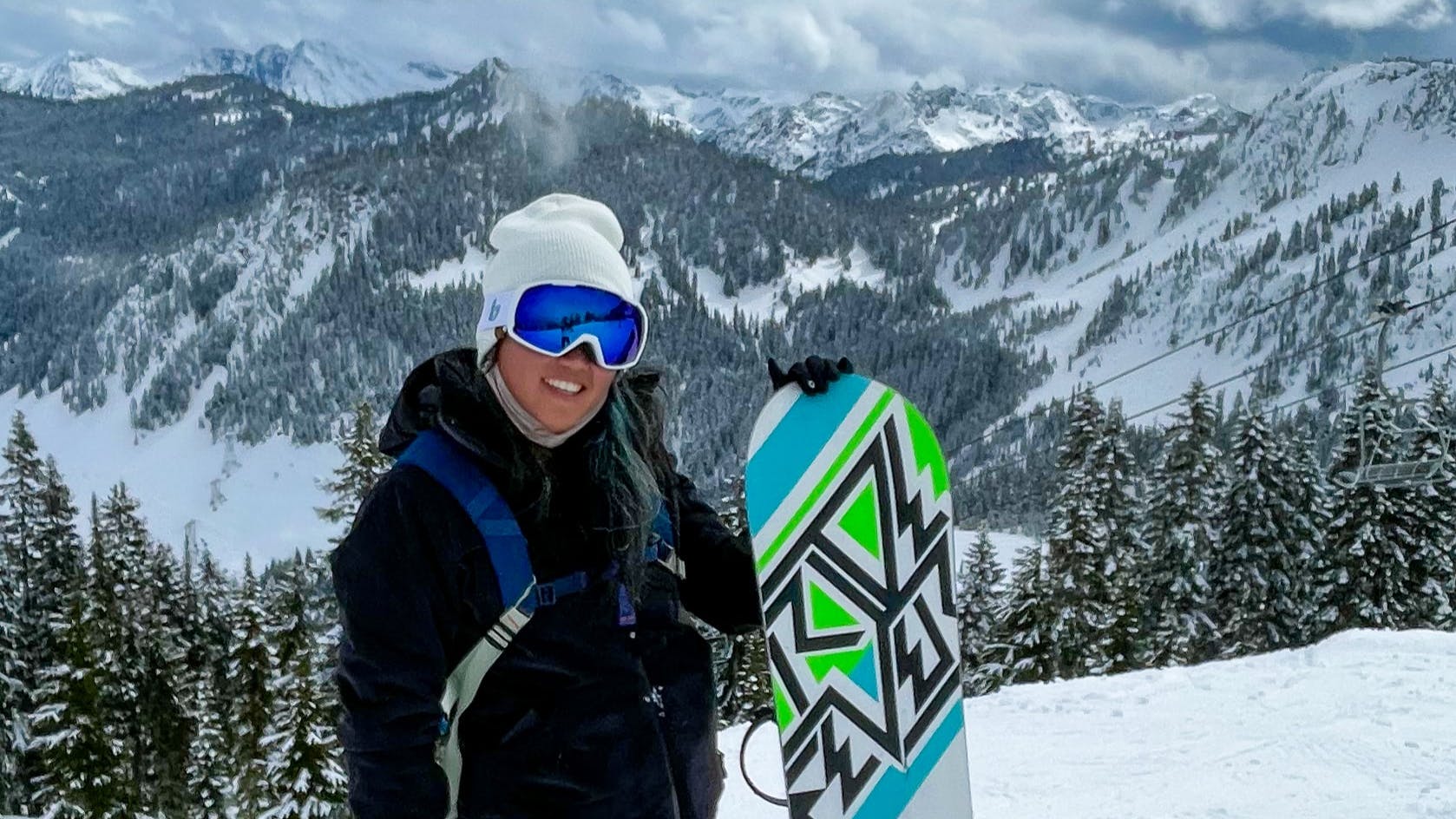 A woman stands holding her snowboard and smiles for the camera. Snowy, tree-covered mountains ruse behind her. 