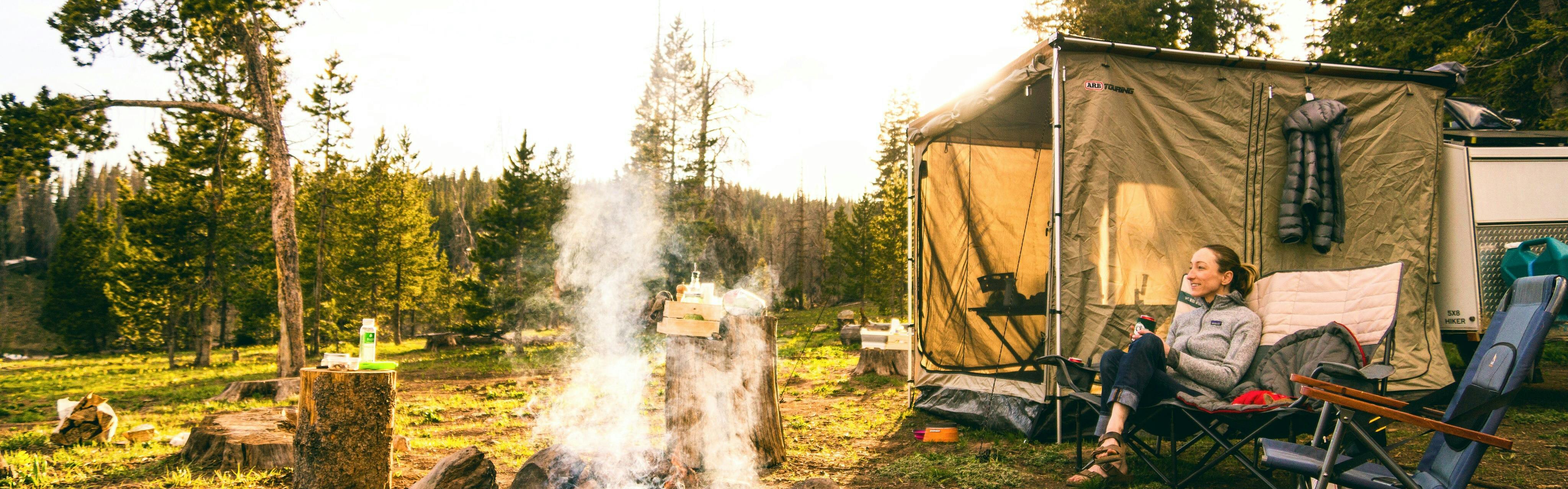A woman sitting by her tent in front of a fire at a campsite in the afternoon.