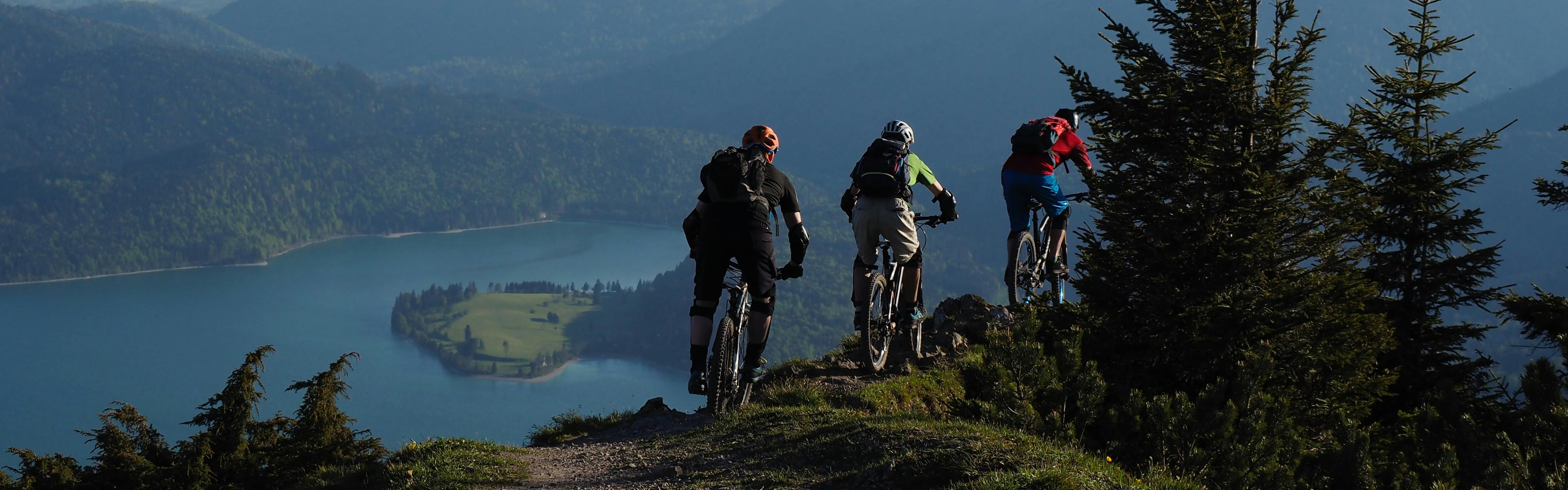 Three mountain bikers ride away from the camera across a narrow ridge. Lakes and hills with trees are in the background to the left.