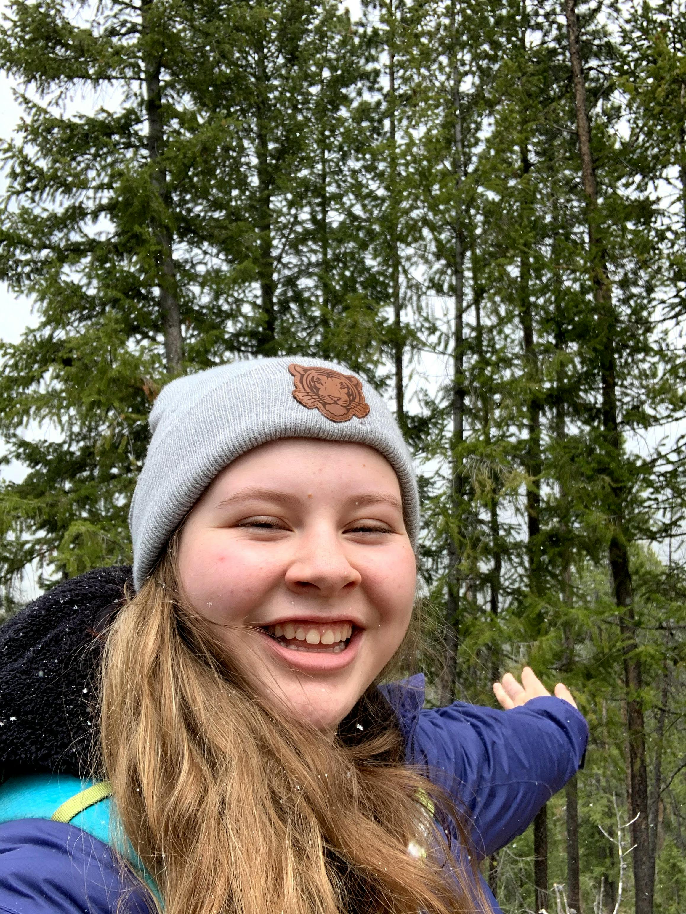 The author takes a selfie amidst tall pines and smiles with a beanie, jacket, rain coat, and backpack on. 