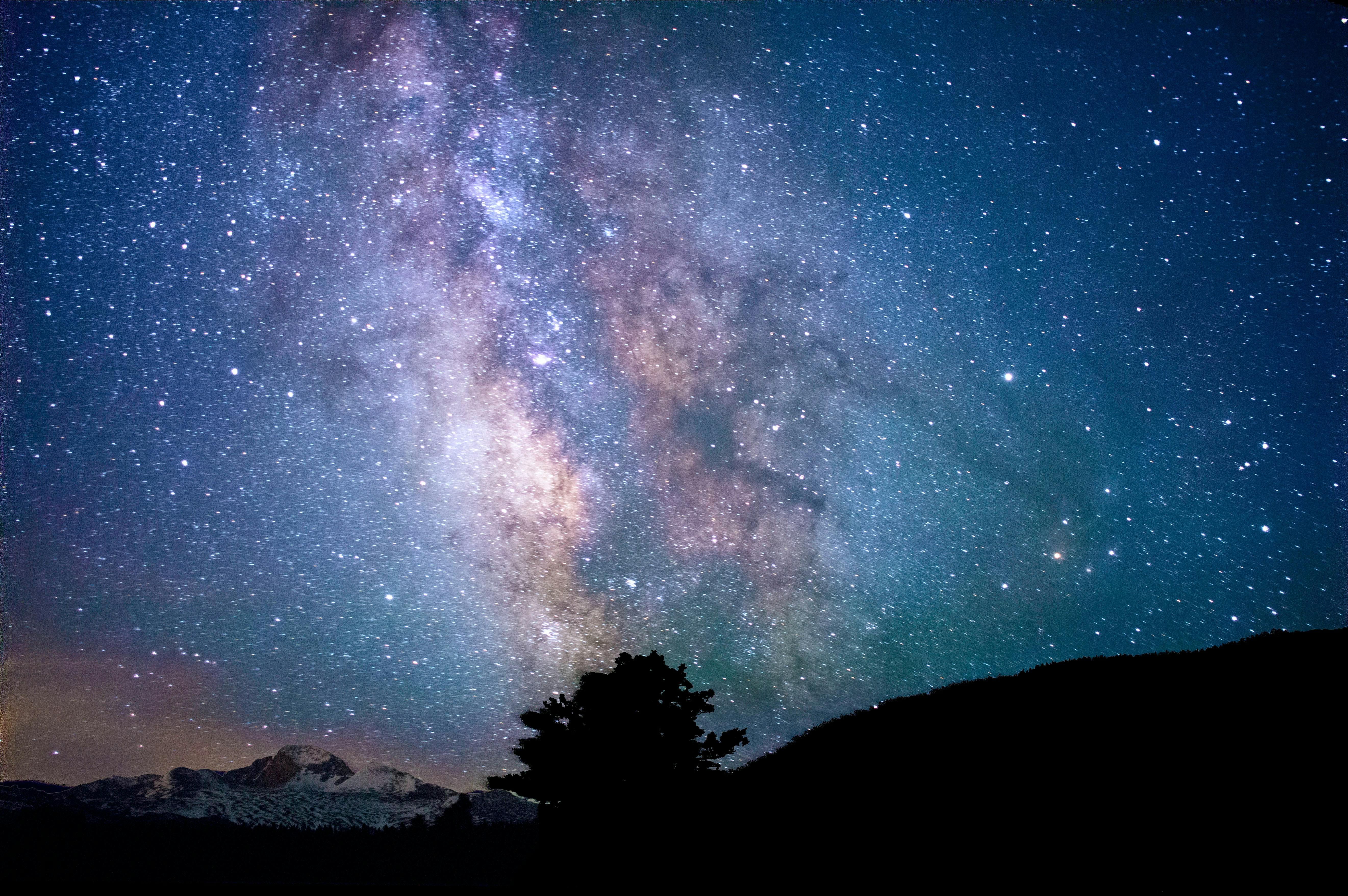 The Milky Way is visible above a dark landscape with a snow-capped mountain rising in the distance. 