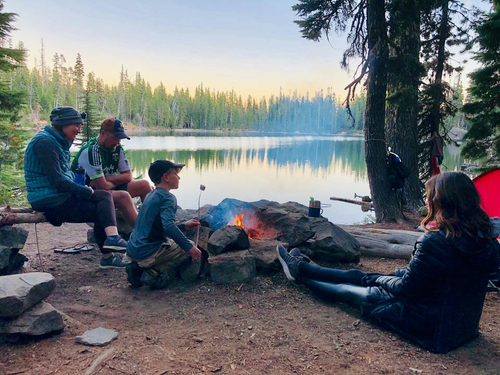 A family around a firepit near a lake. The boy is roasting a marshmellow.