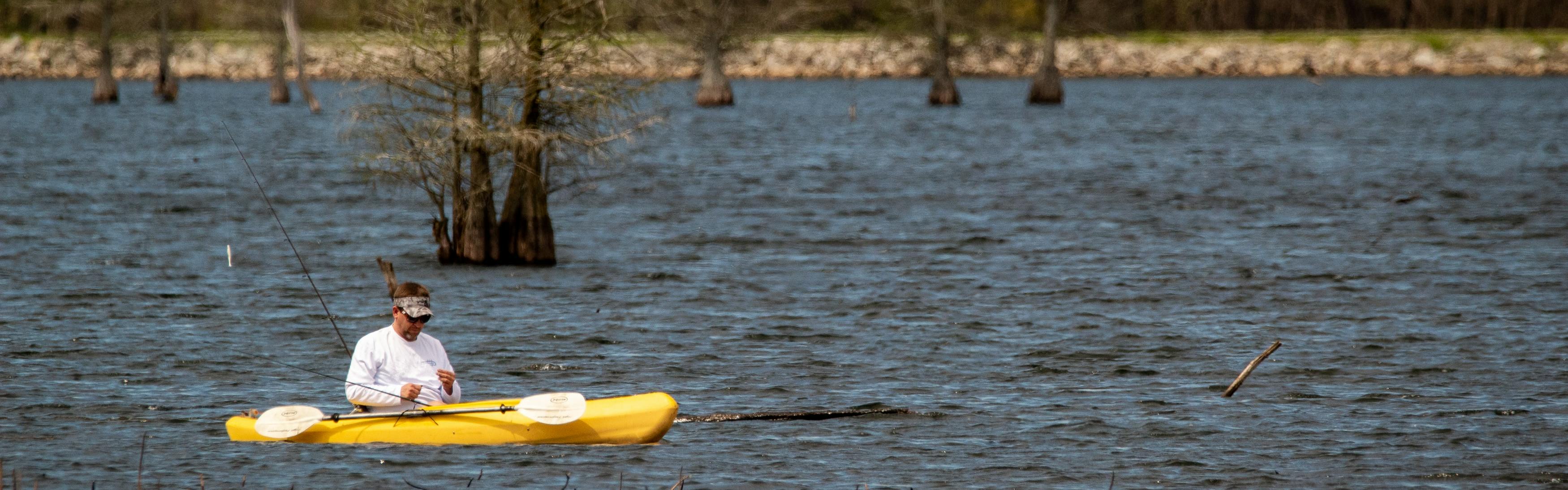 A man sits in a yellow kayak low to the water and fiddles with some piece of gear. His fishing rod points out of the boat behind him and his paddle is next to him in the boat. 