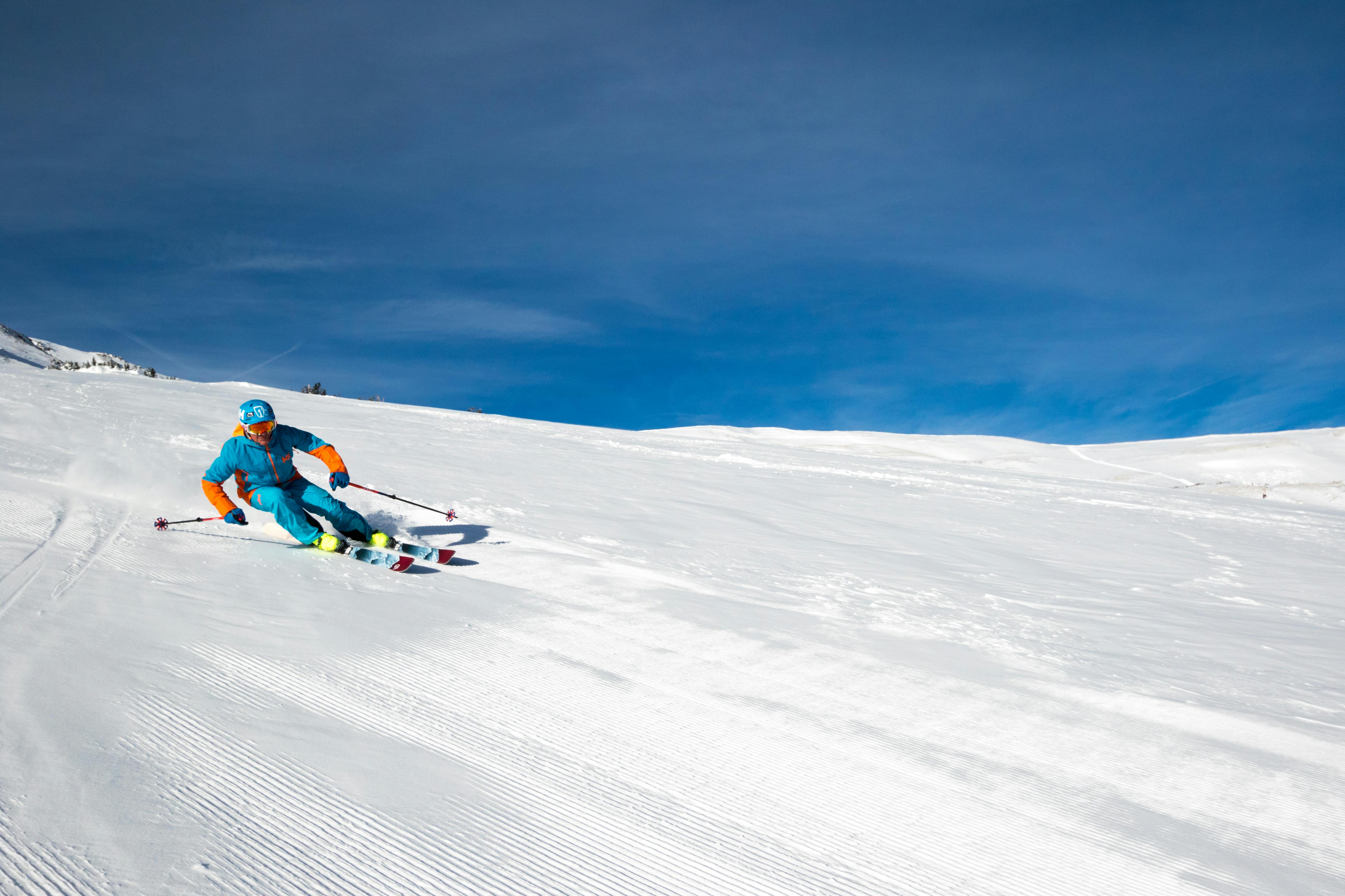 A man in a blue and orange ski jacket and pants skiing fast down a steep groomer slope