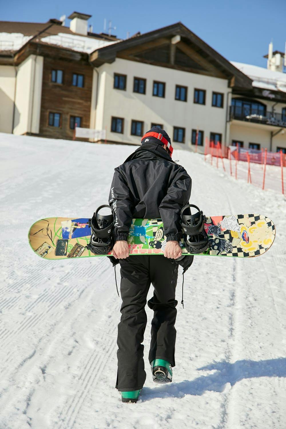 Man walks up a snowy hill towards the lodge. He is carrying a snowboard behind him and also looks to be stretching a bit. 