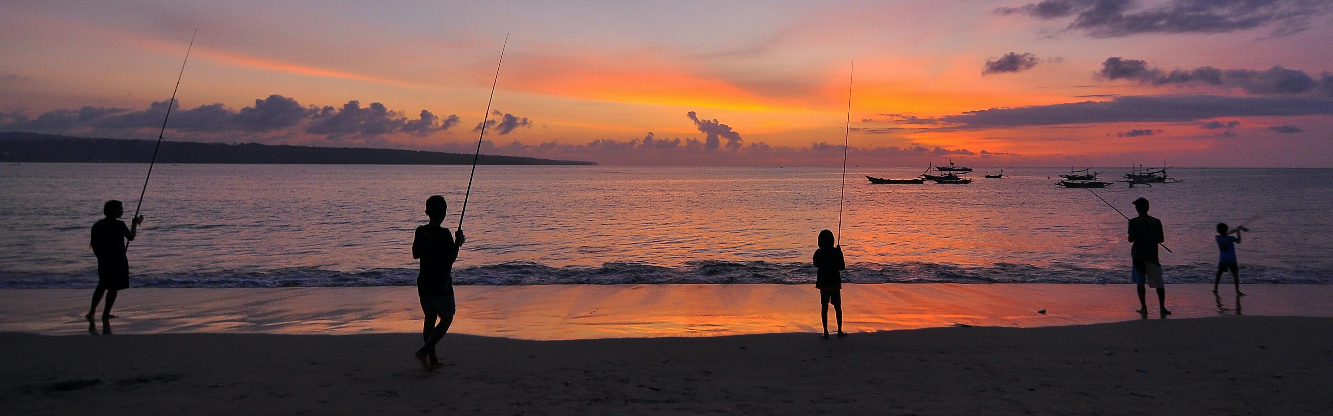 Several people fish from the shore of the ocean as the sun sets. 