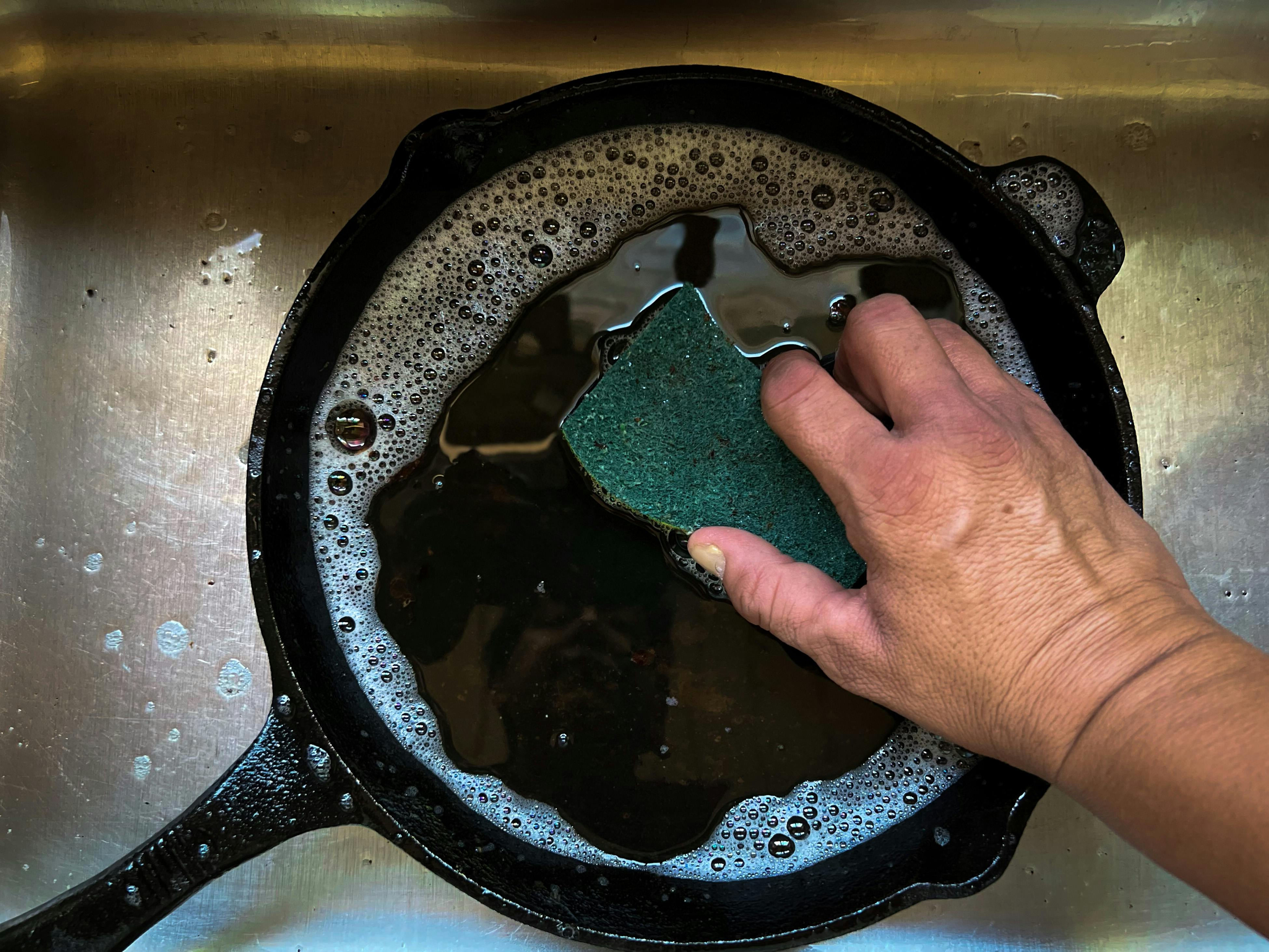 The author washes his cast-iron pan in the sink with a sponge.