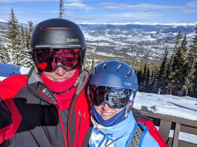 Two skiers sitting on a chairlift wearing helmets and goggles. 