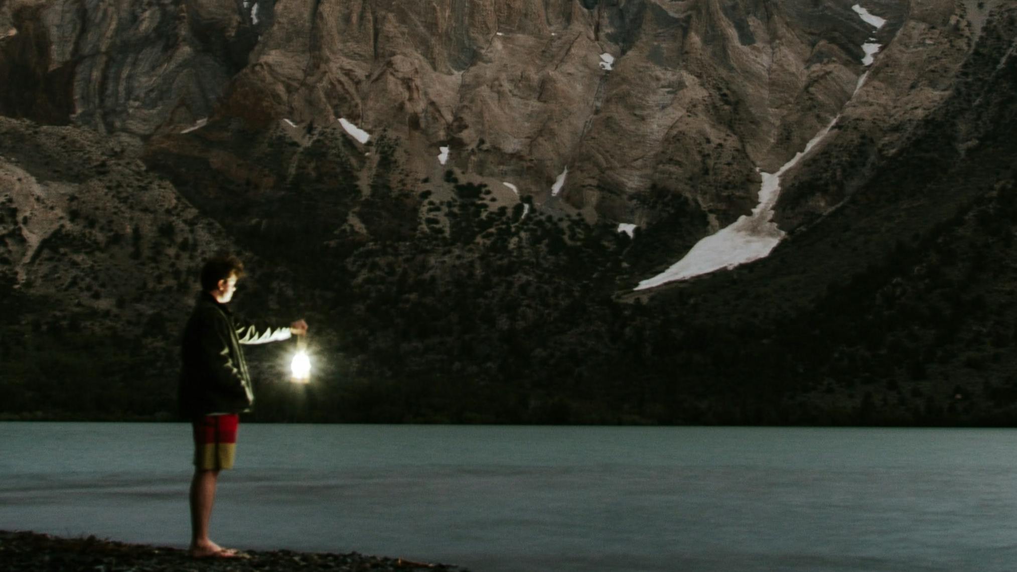 A person standing on the edge of a lake holding up a lantern. There is a mountain and a stream in the background and it is starting to get dark out. 