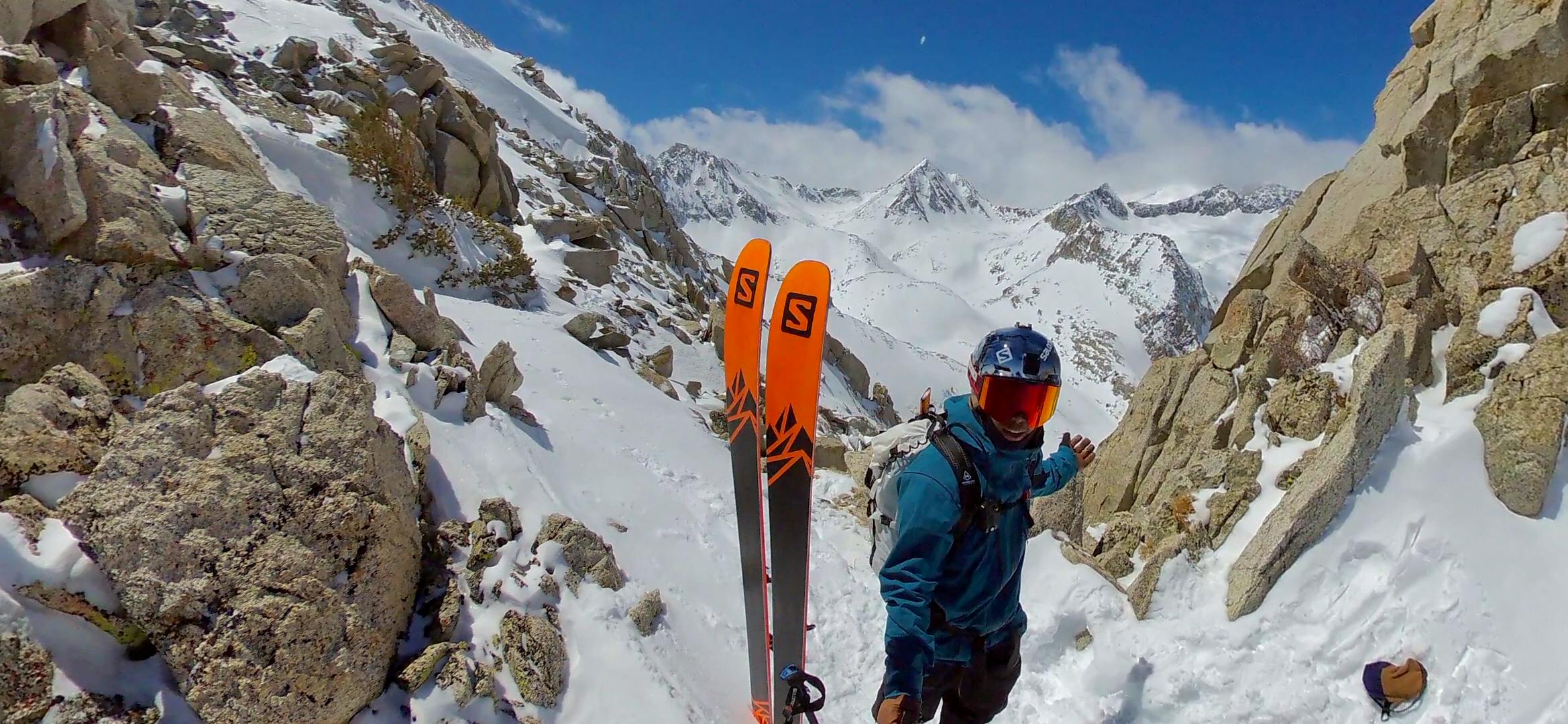 Josh takes a selfie on the mountain while wearing a google and a helmet. His Salomon skis are propped up next to him.