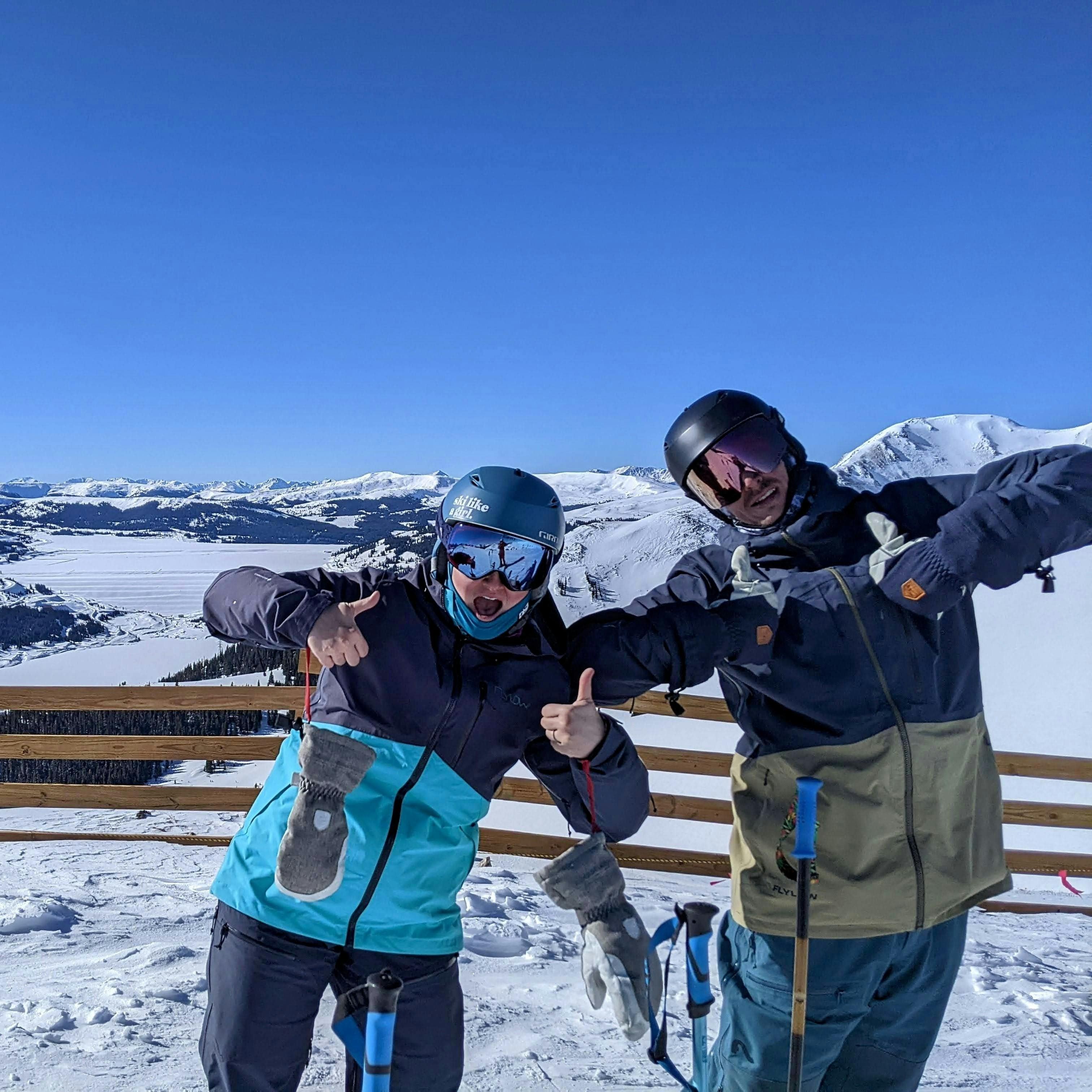 Two skiers standing at the top of a ski run giving a thumbs up to the camera. 