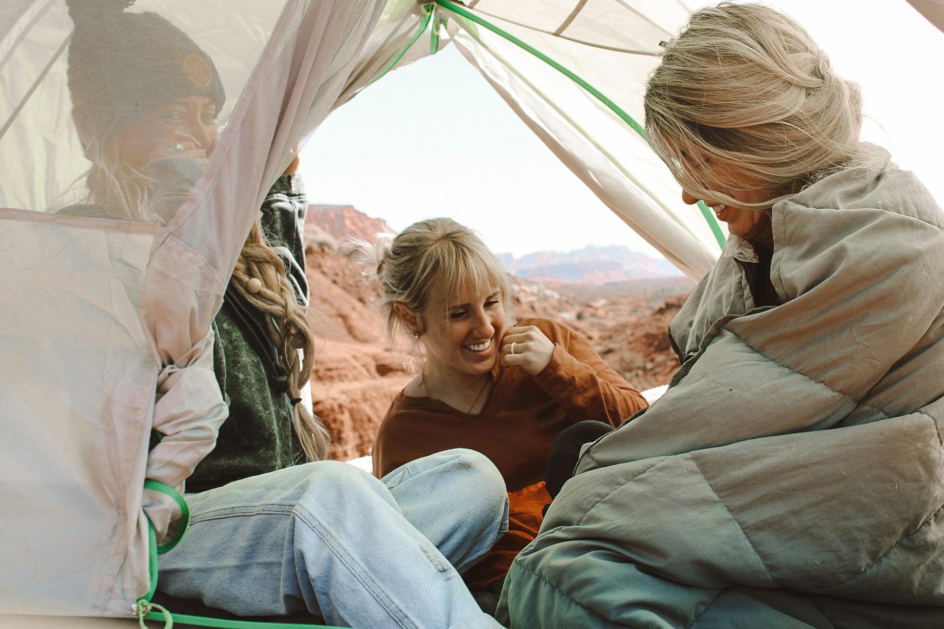 Three girls sit in a tent laughing. There are blankets in the tent and one person is wearing a blanket around their shoulders. 