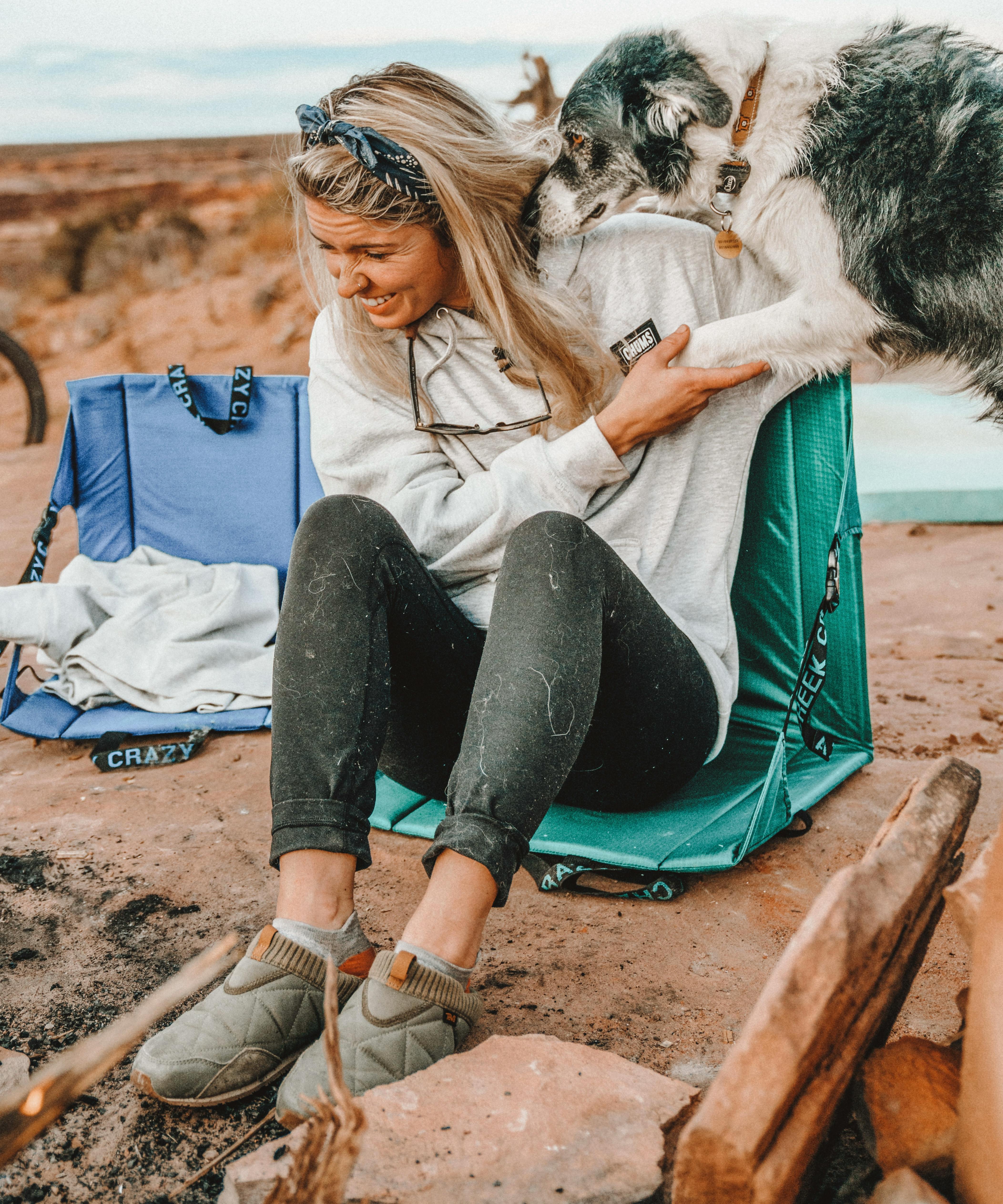 A dog jumps on a woman sitting in a chair around a campfire.