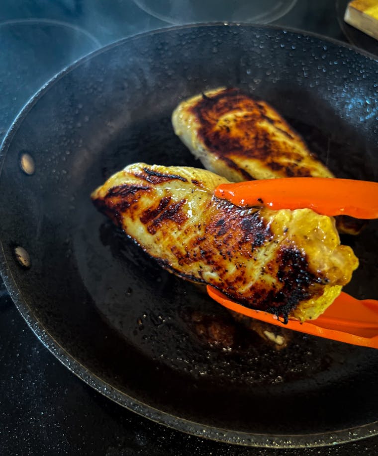 A pair of tongs lifting up a chicken breast from a pan. 