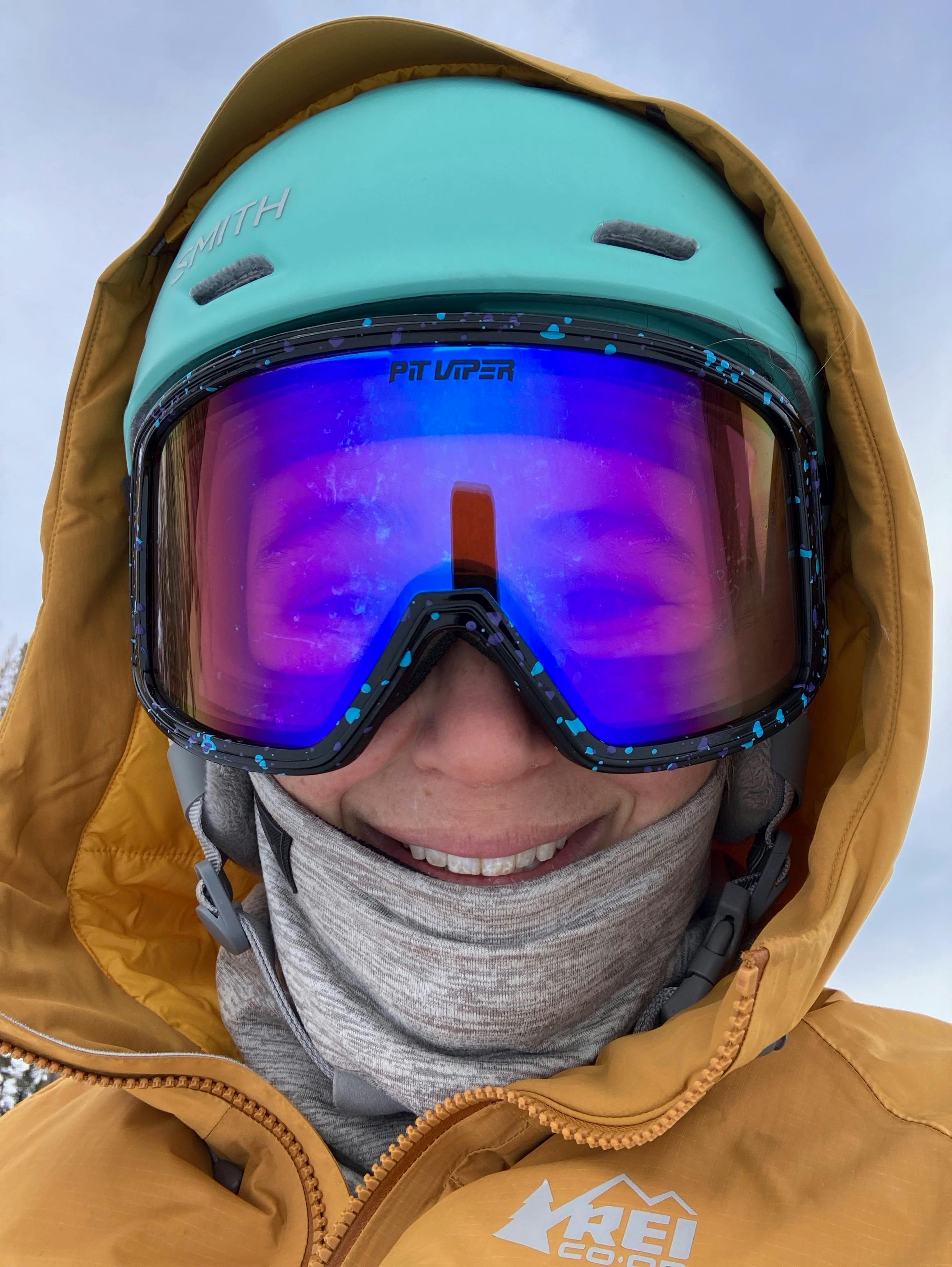 Close up of a snowboarder's face with pale skin, a light blue helmet and blue and purple mirrored lenses smiling into the camera.