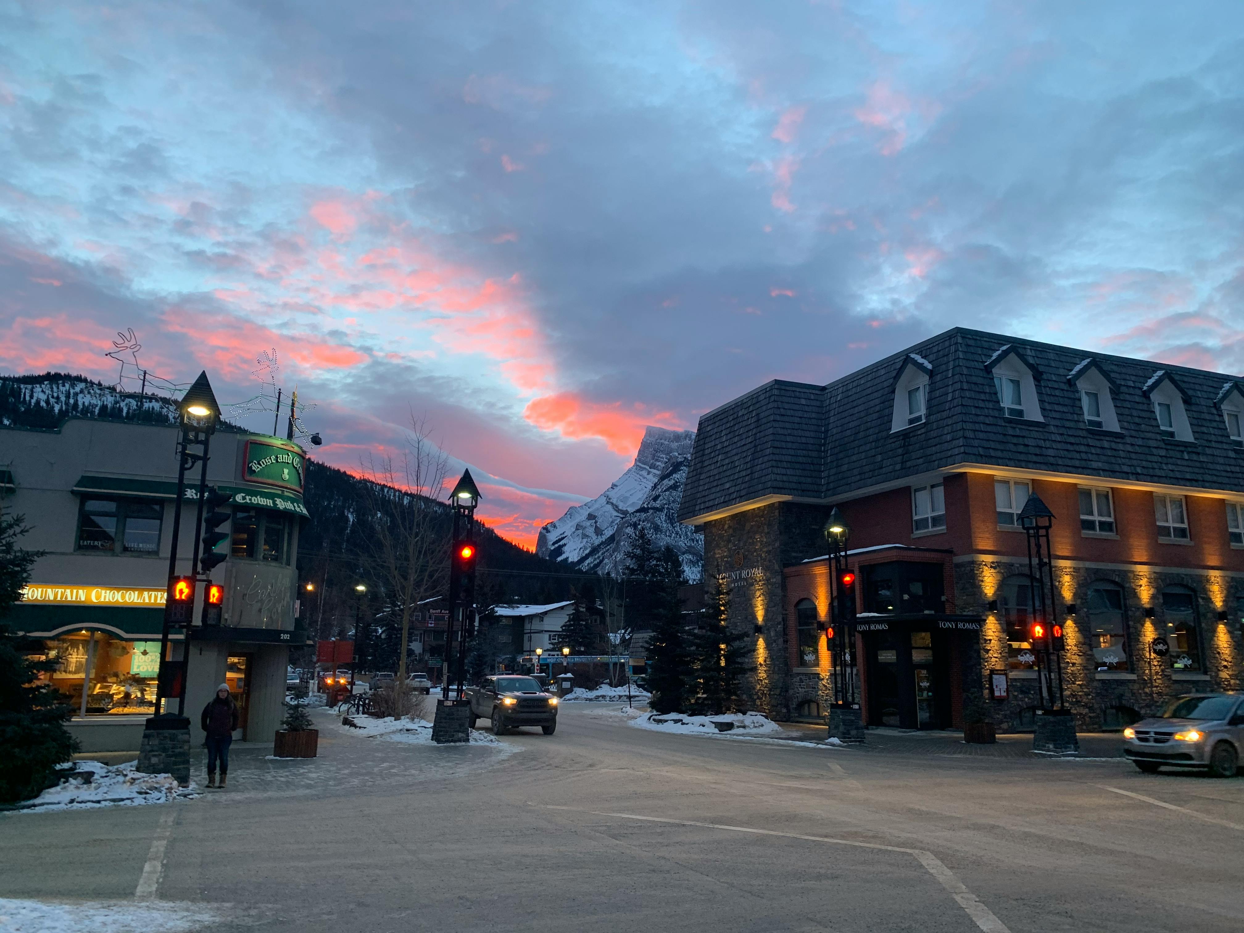 An image taken on a street corner in Downtown Banff. In the background, a jagged, snow-covered peak rises about the town, and the sky glows pink and blue in the cloudy sunset.