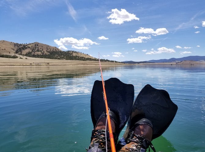 An image of flyfishing boots and someone's orange rod sticking out of a lake with a blue sky above. 