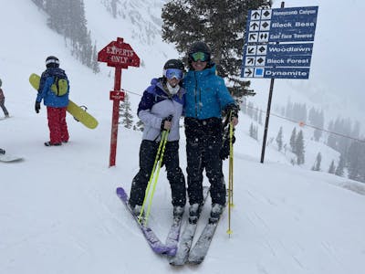 Two skiers stand on a ski run. One has his arm around the other and there is a trail sign behind them.
