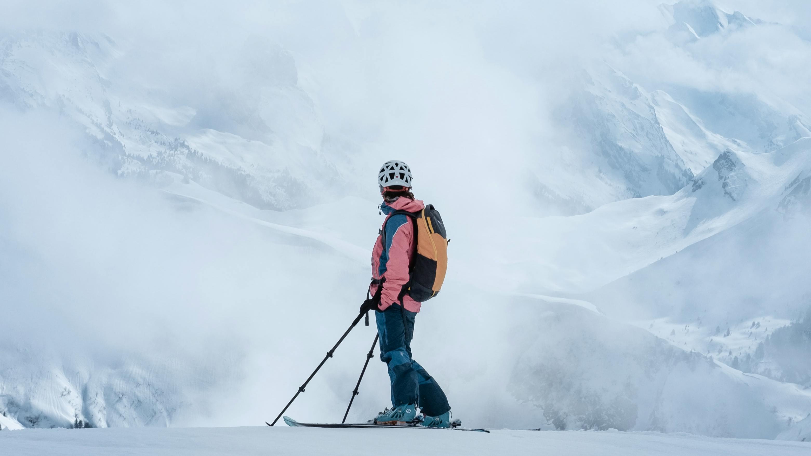 Someone stands sideways to the camera with their head looking away and towards snow-covered peaks that are covered in fog. They're outfitted for a backcountry tour.
