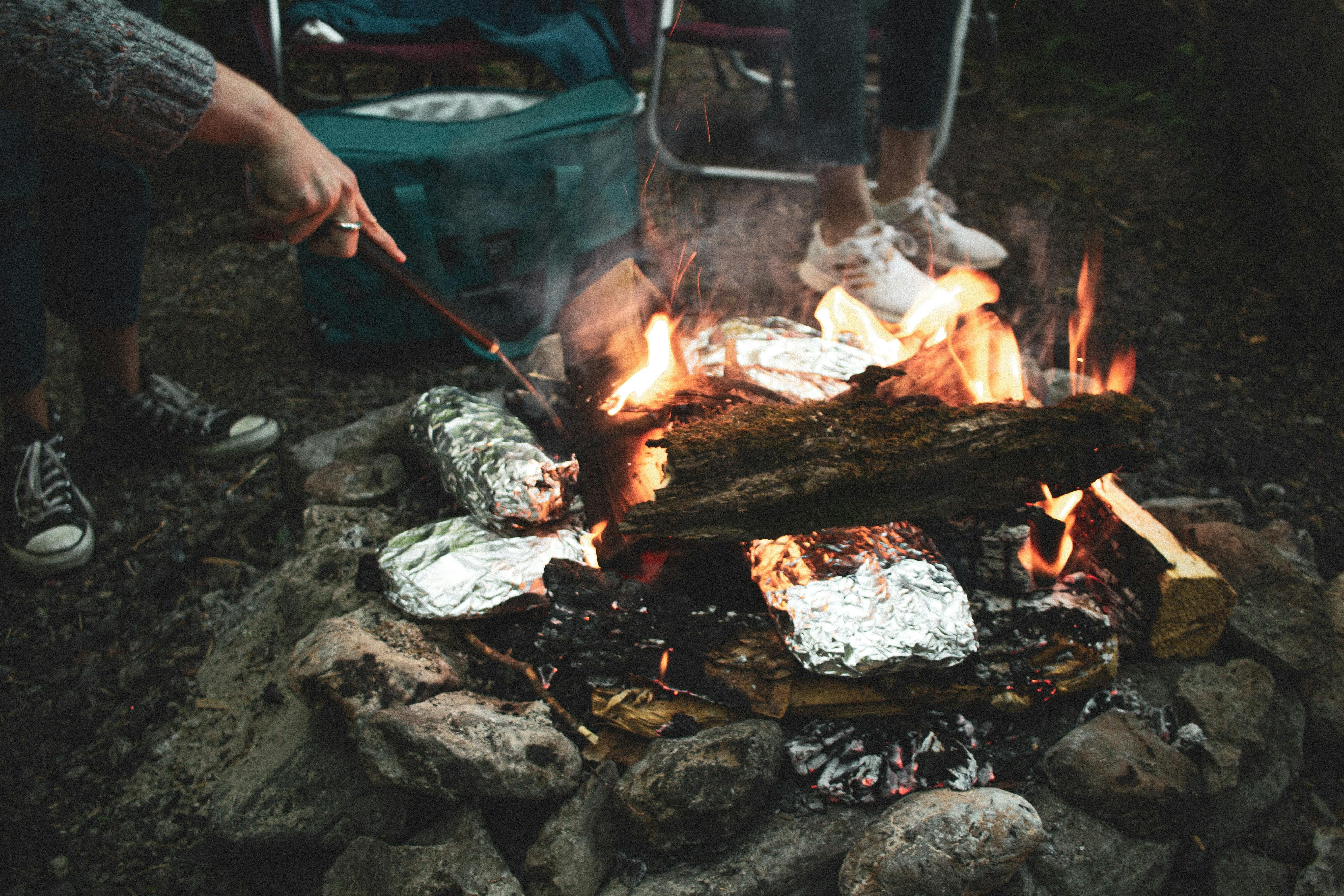 Someone pokes at food wrapped in tinfoil that sits between logs in a campfire. 