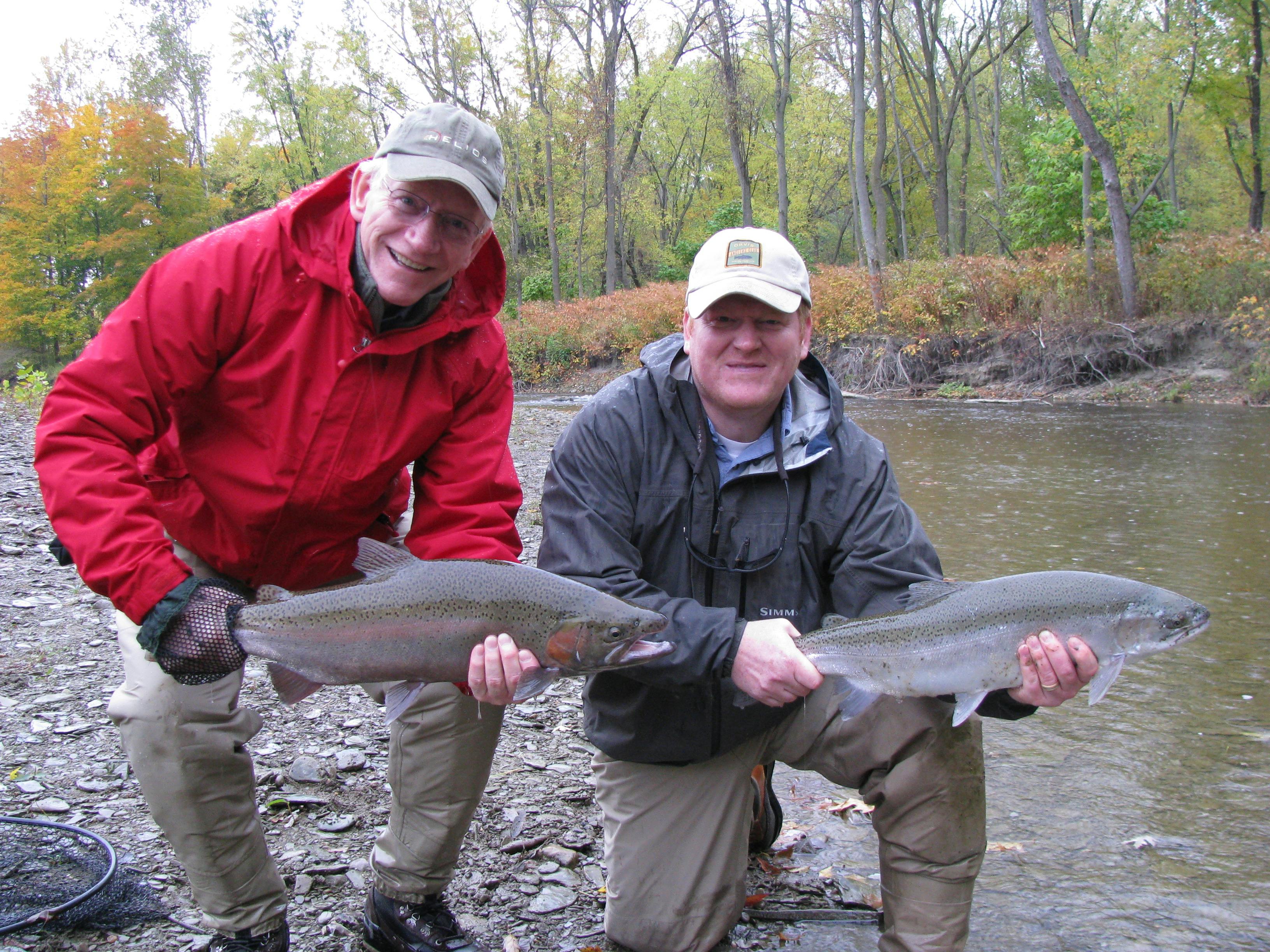 Two men squat next to a river while holding out their catches and smiling. 