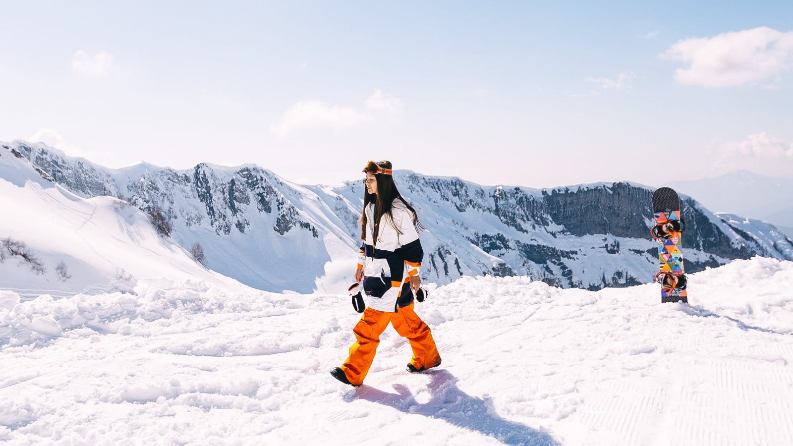 A woman walking away from her snowboard. She is wearing snowboard gear and at the top of a snowy mountain. 