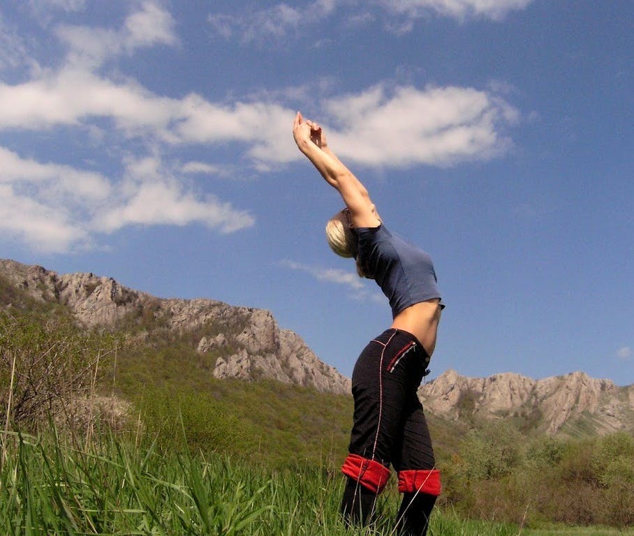 A woman stretches in a field. There are some mountains in the background.
