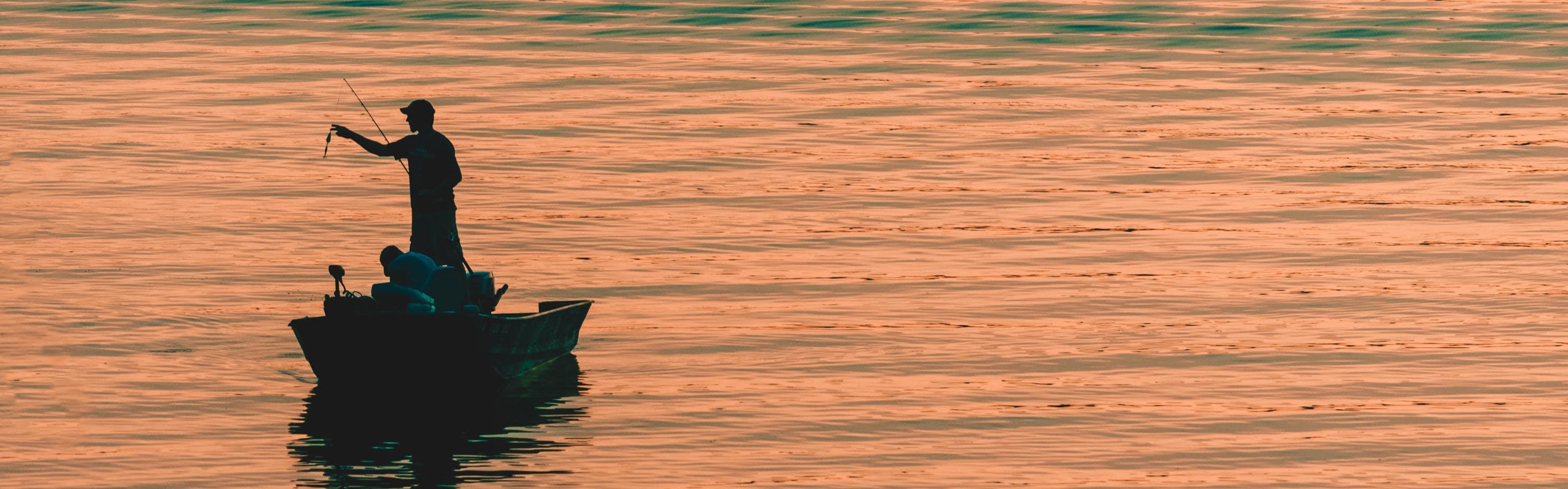 Someone preps their line while standing in a boat. The scene is silhouetted against a lake full of ripples and peach-colored in the setting sun.