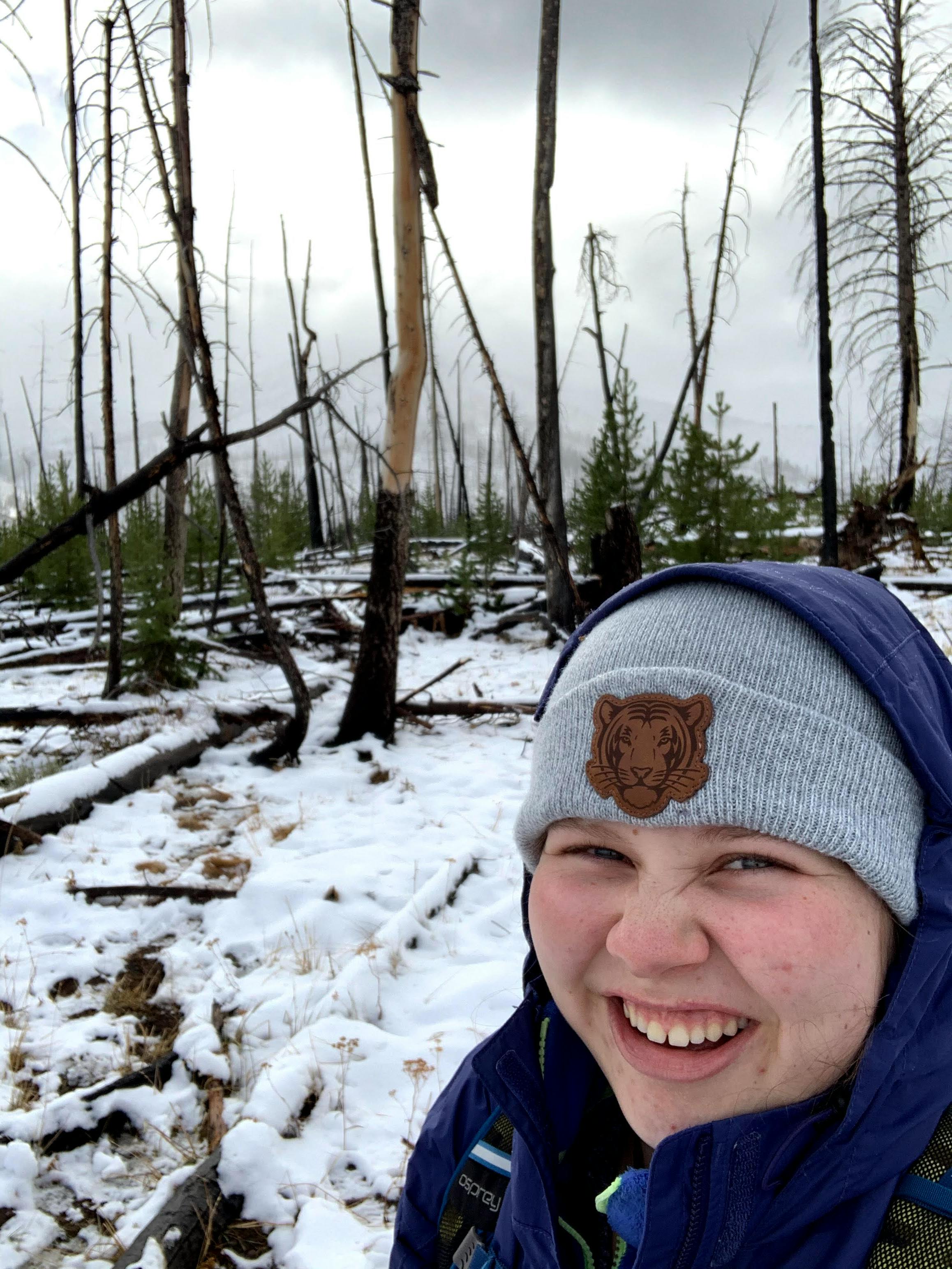 The author smiles in a beanie and a hood with fire-burned trees in the background.