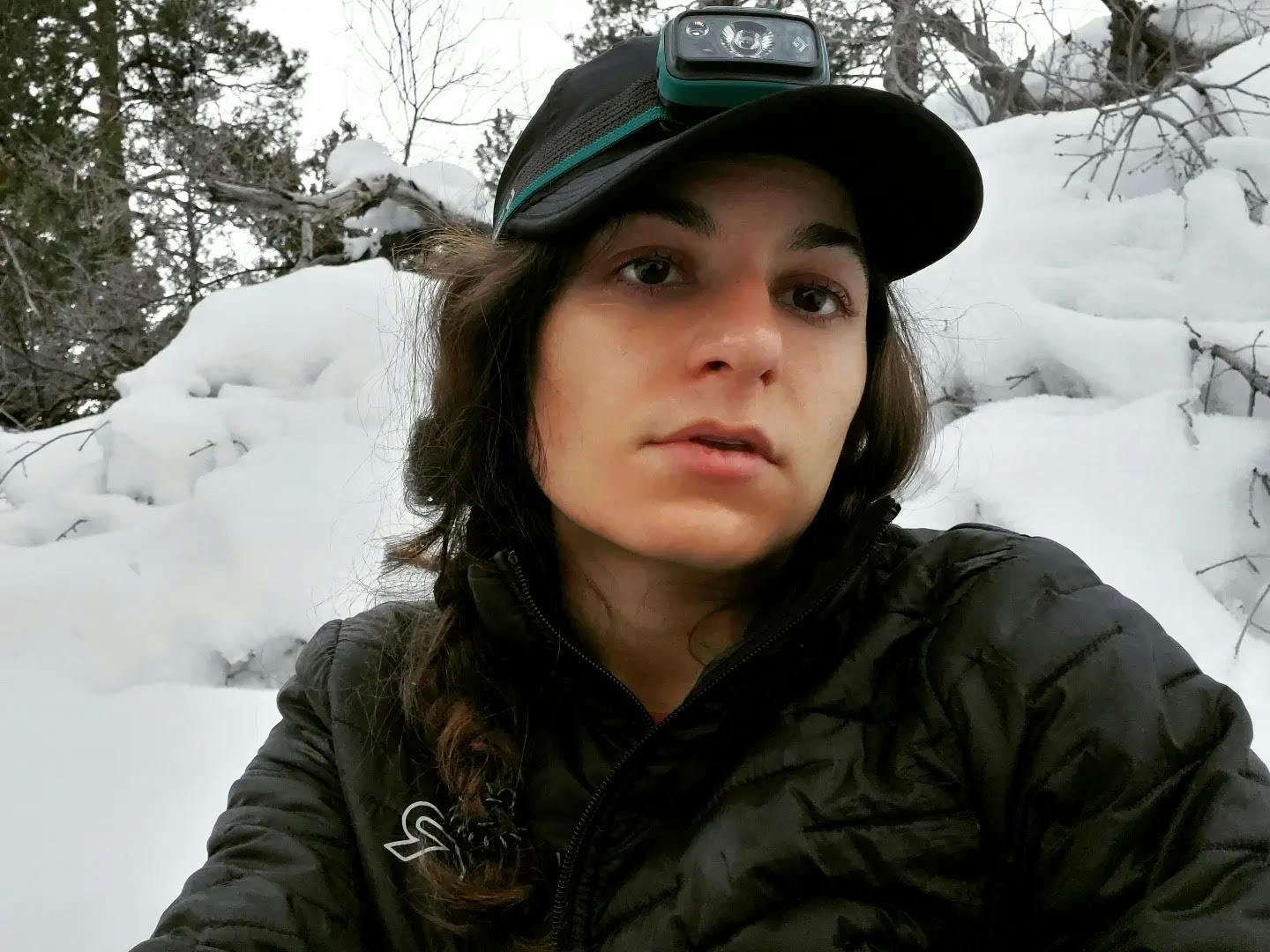 A selfie of a dazed looking hiker wearing a headlamp and hat. She is sitting in a snowy area.