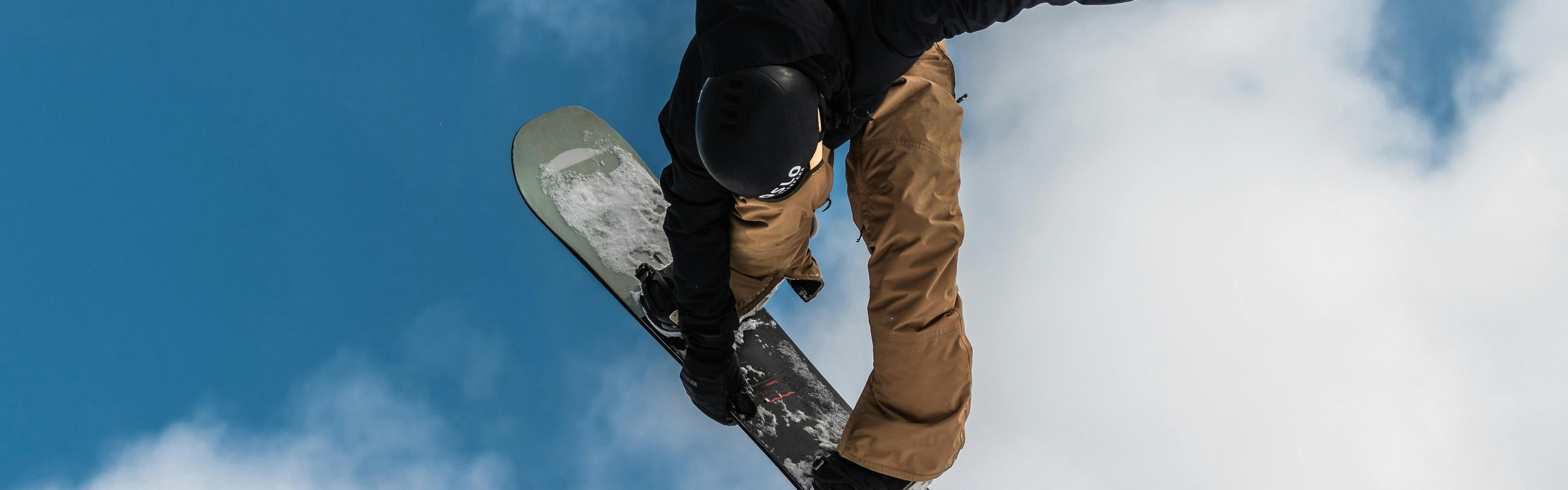 A snowboarder in a black jacket and brown pants flying through the air against the blue sky holding onto his snowboard while executing a jump trick.